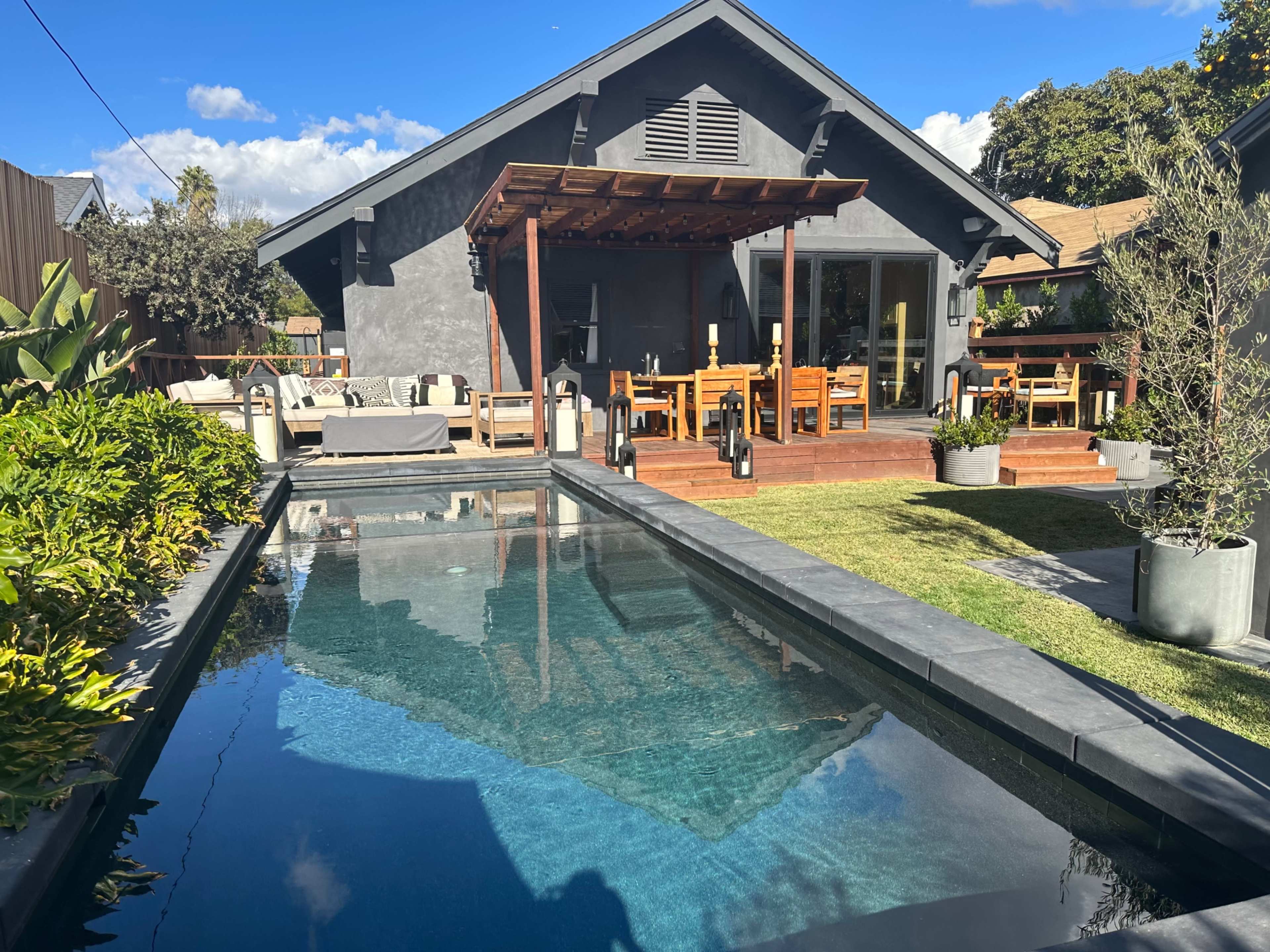 A modern backyard features a swimming pool, wooden deck, and dining area alongside a gray house.
