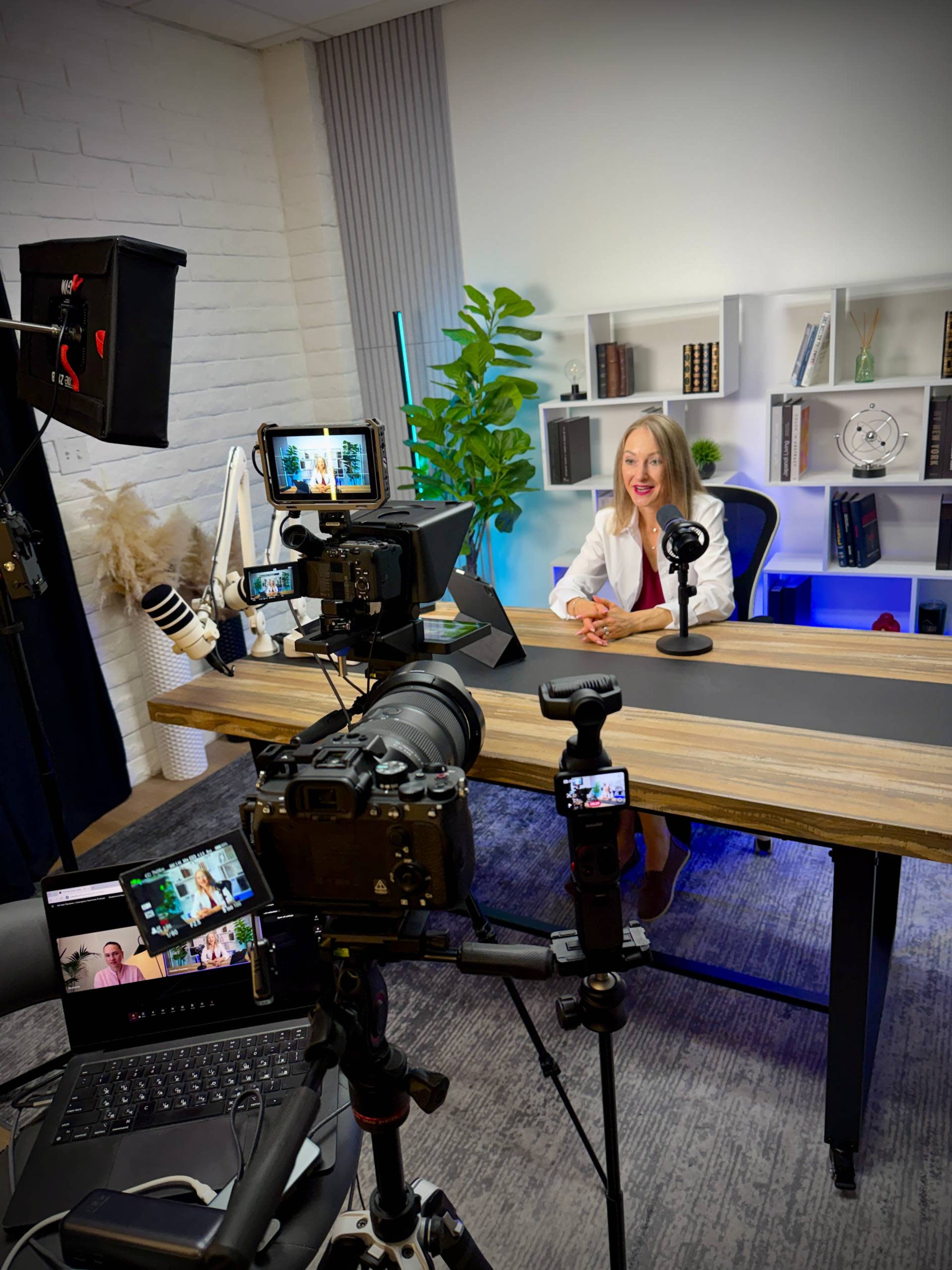 A woman in a white coat sits at a desk with multiple cameras and microphones set up for filming in a well-lit, modern studio.