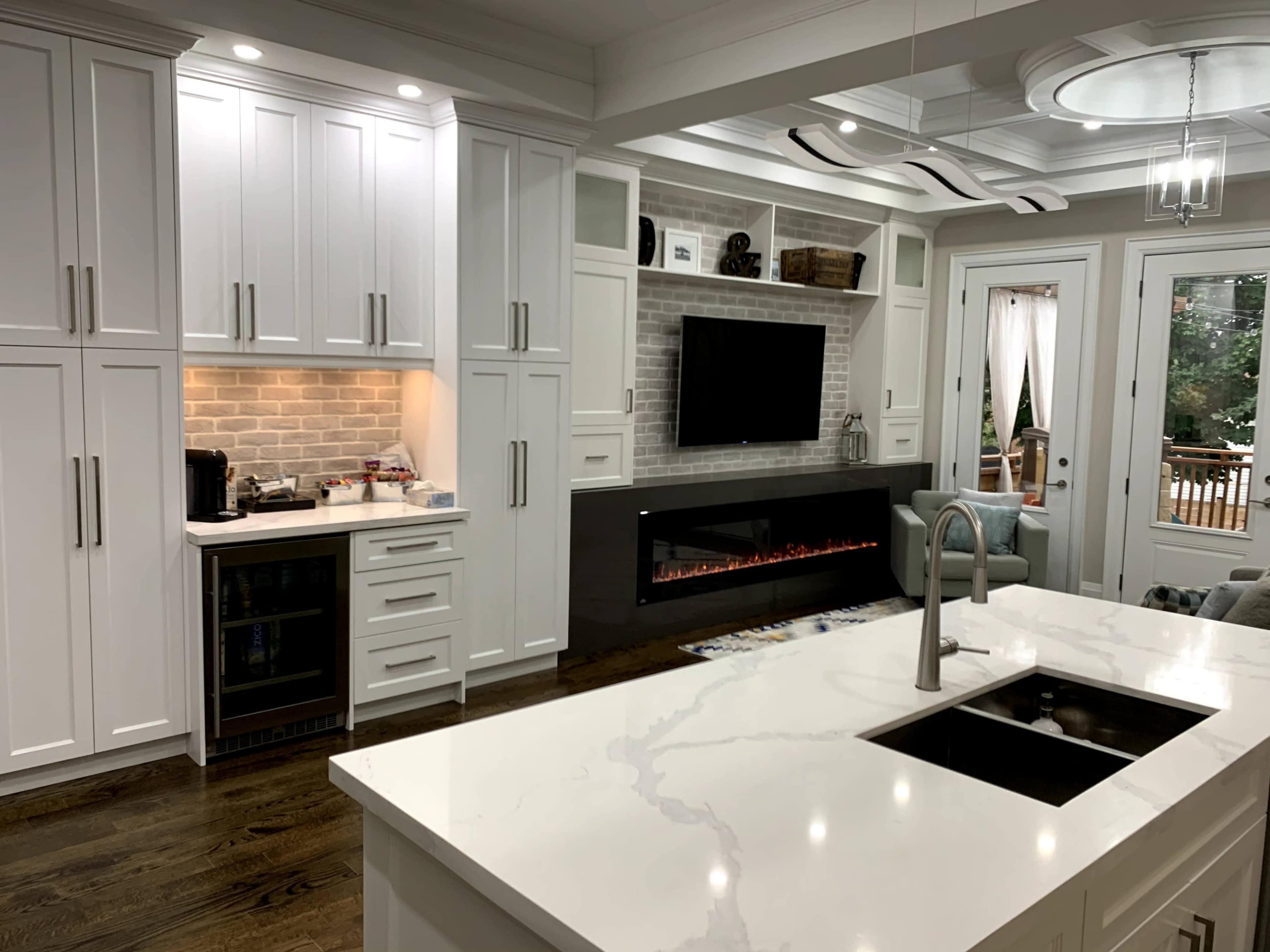 A modern kitchen with white cabinets, a large island with a black sink, and a wall-mounted television above an electric fireplace.
