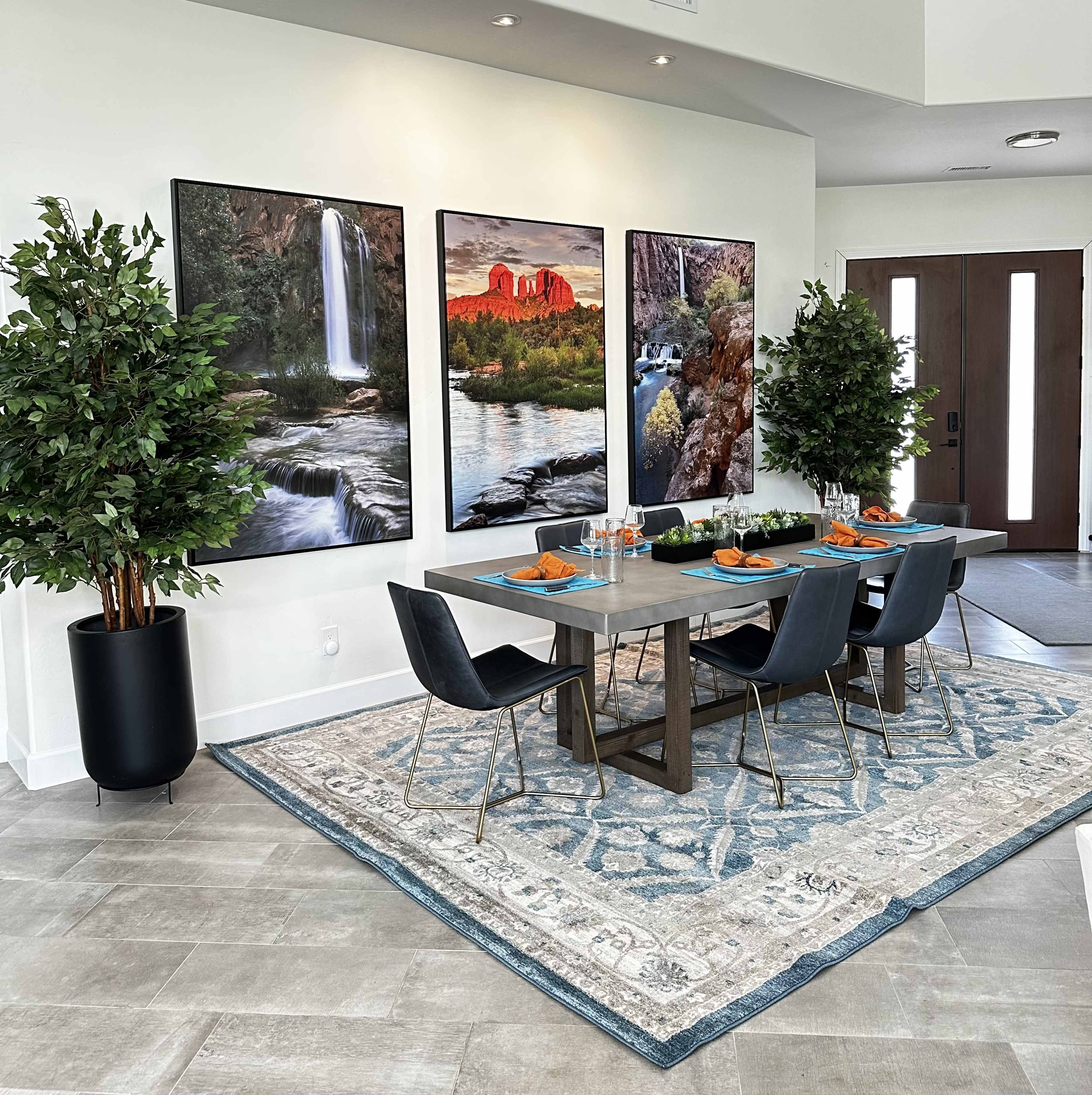 A modern dining area features a table set for four, flanked by two green plants, with large landscape photographs on the wall and a decorative rug beneath.