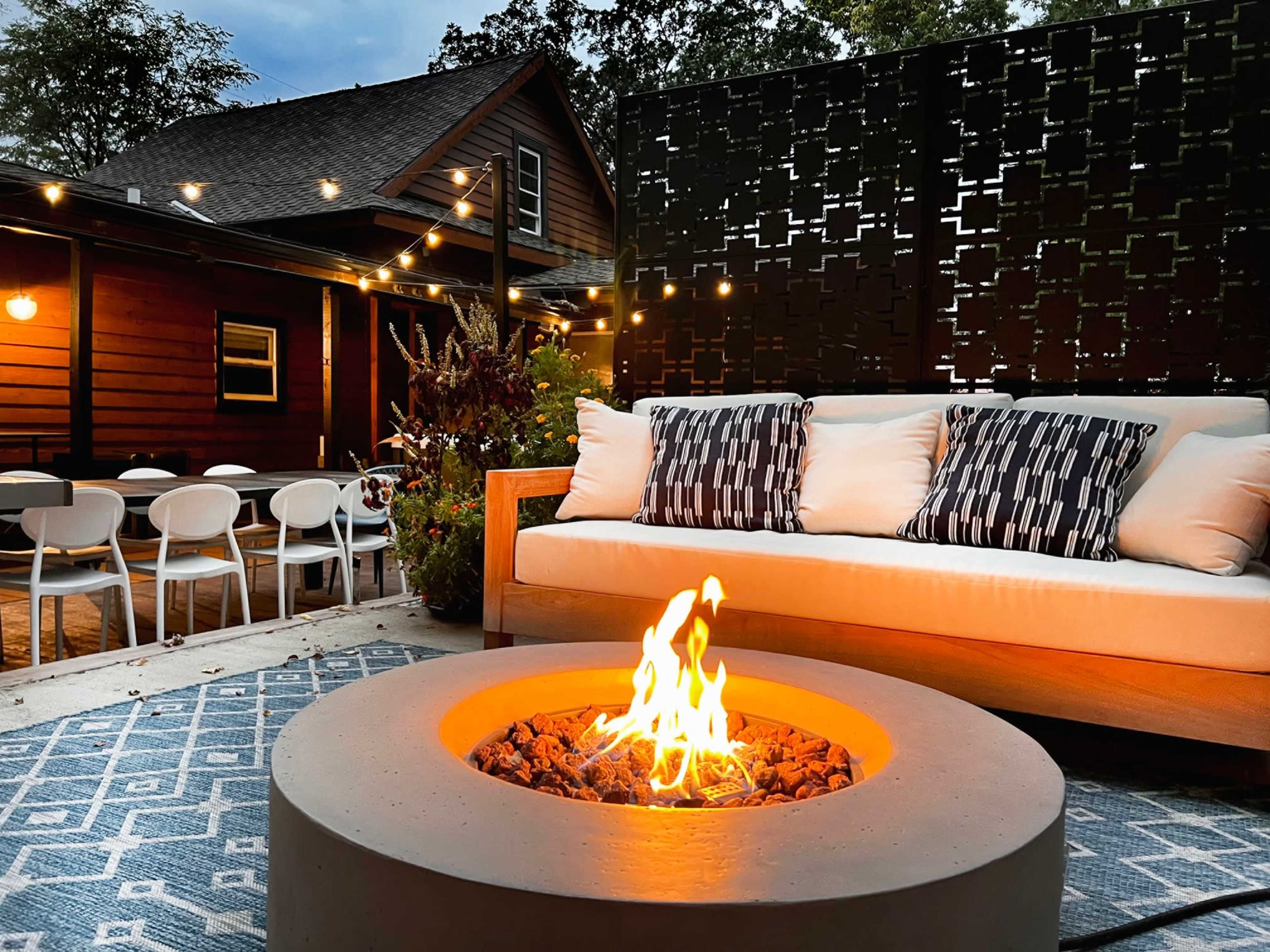 A modern outdoor seating area features a circular fire pit surrounded by a couch with patterned cushions, set against a backdrop of a wooden house and string lights.