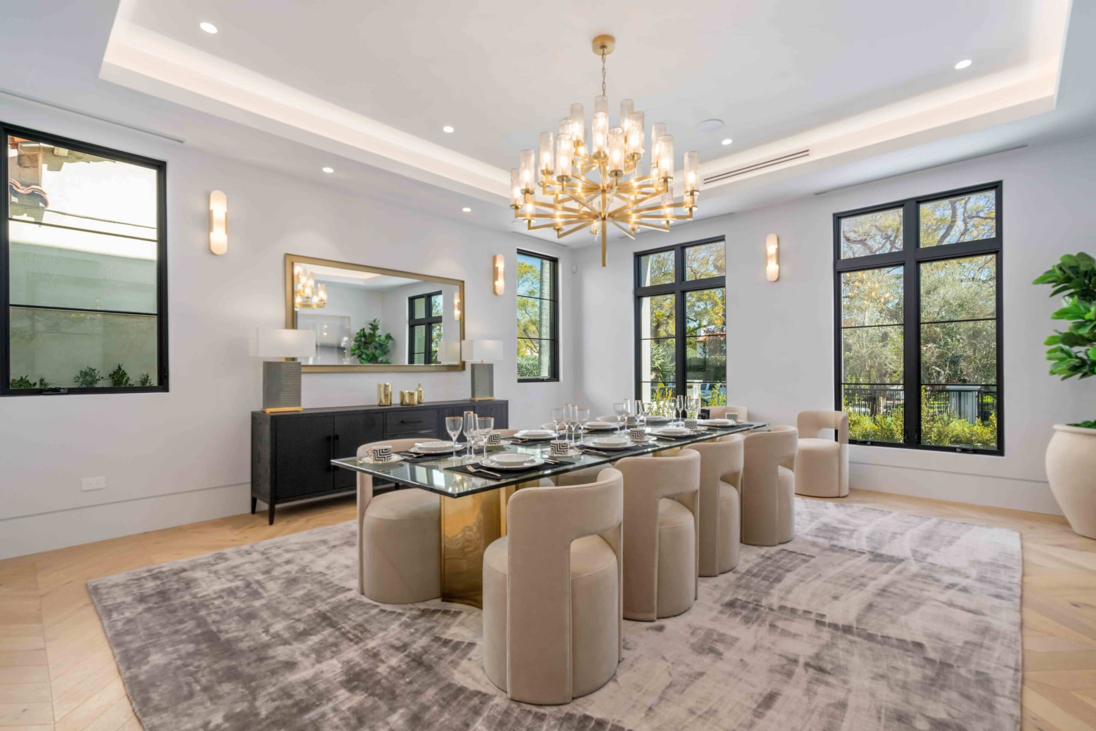The image shows a modern dining room featuring a large glass dining table surrounded by upholstered chairs, with a decorative chandelier and large windows allowing natural light in.