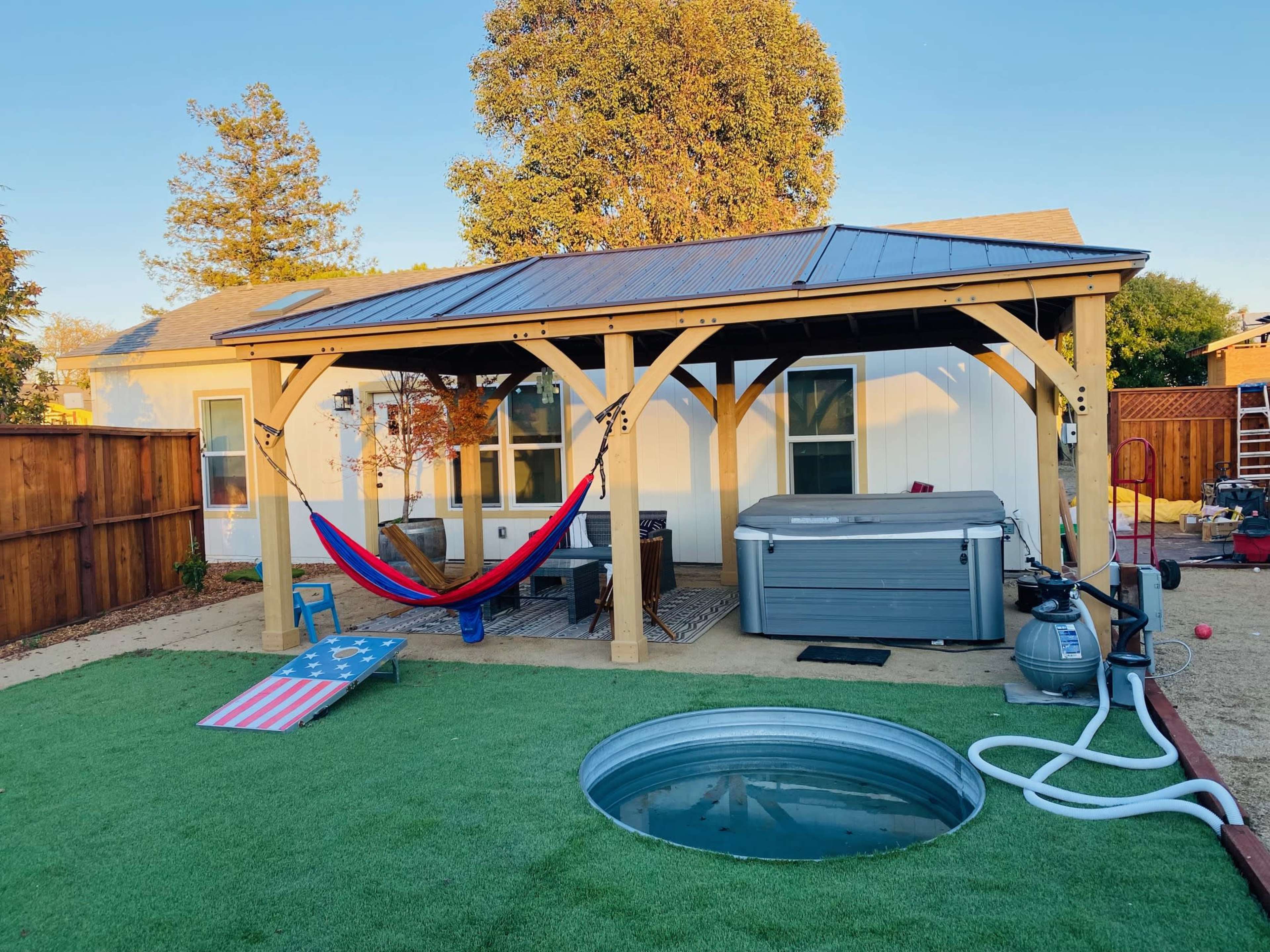 A wooden pergola with a hammock and a hot tub is situated in a yard featuring artificial grass and a metal fire pit.