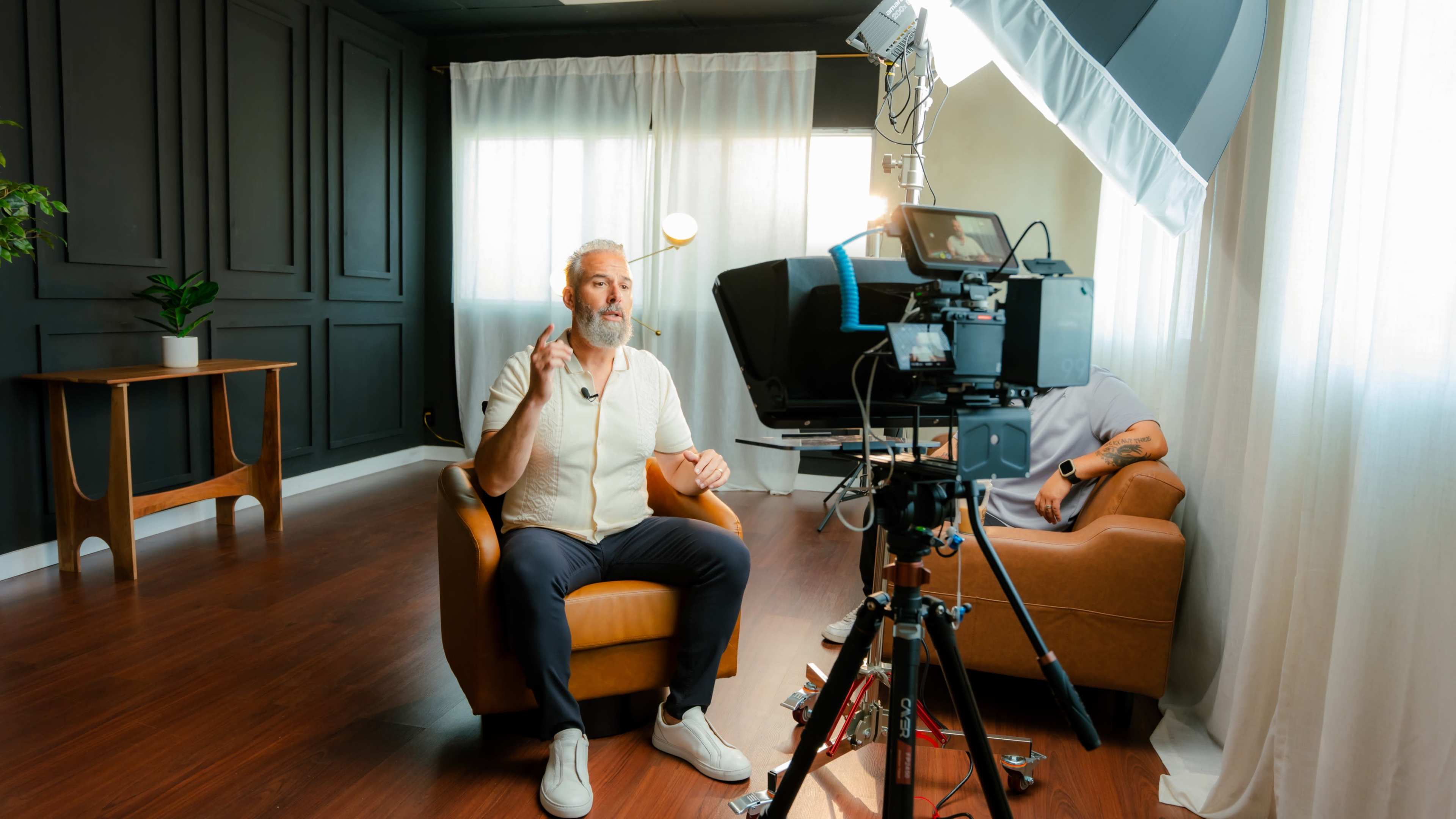 A man with a beard speaks while sitting in a leather chair next to a camera setup in a well-lit room.