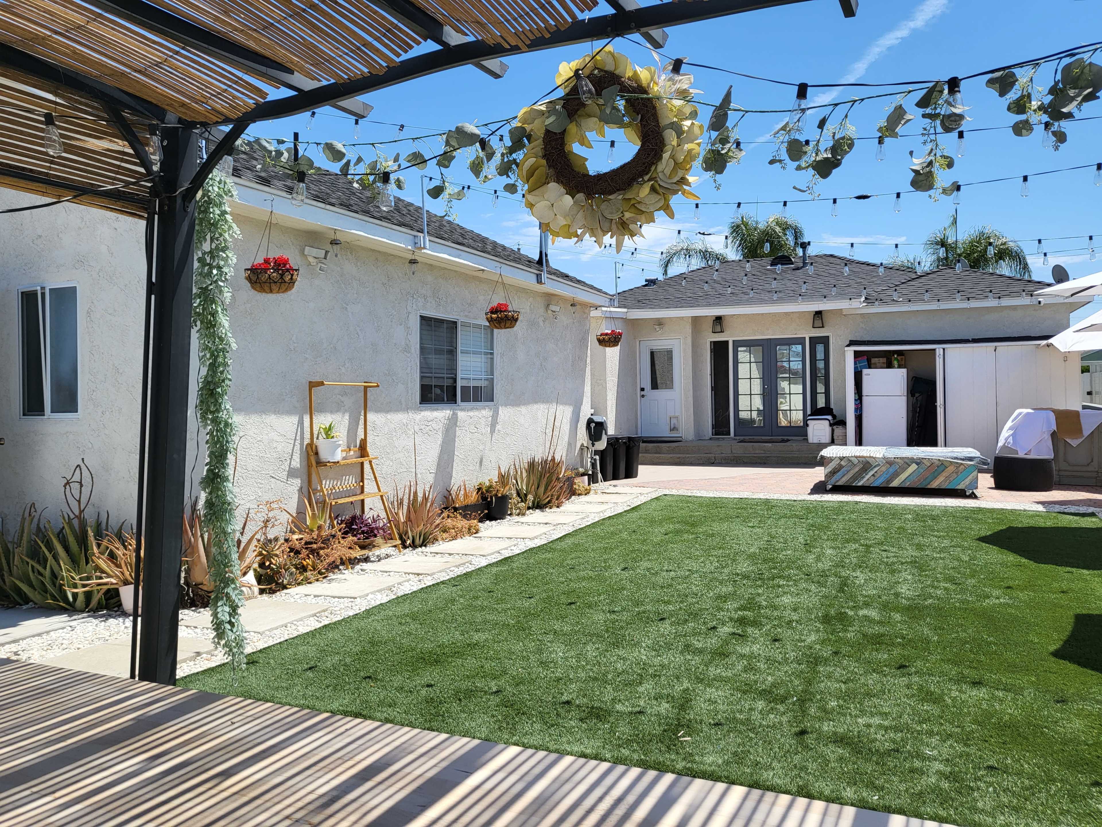 A backyard with artificial grass, a covered seating area, and potted plants hanging from a pergola.