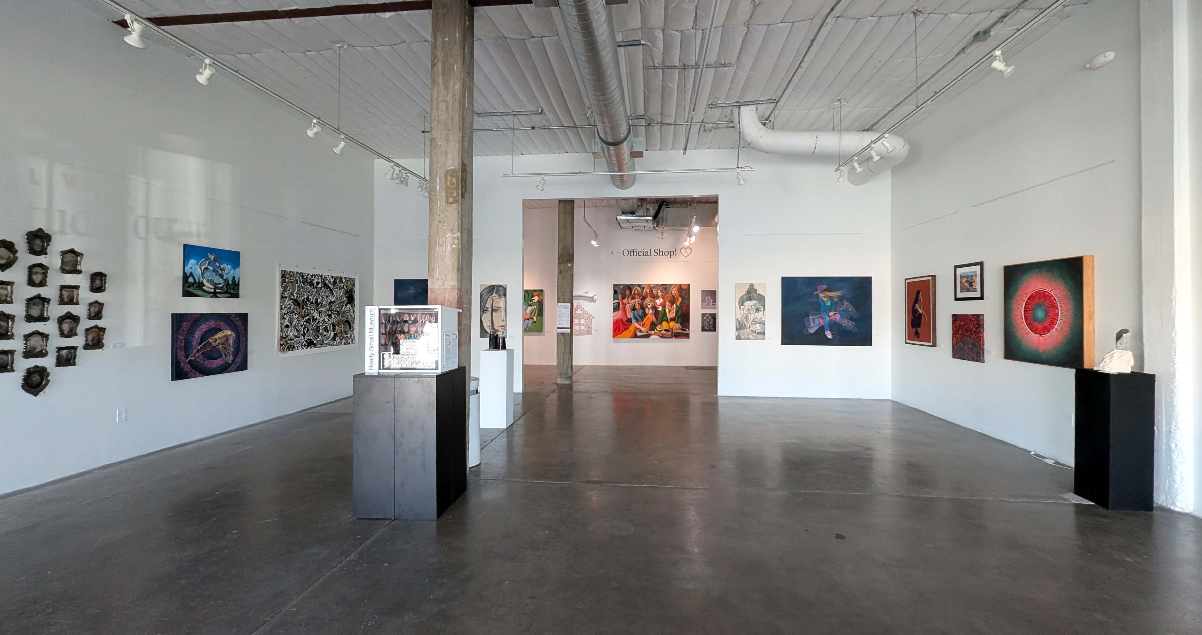 The image shows an art gallery with white walls, various framed artworks displayed along the sides, and a central area featuring sculptures and more art pieces.