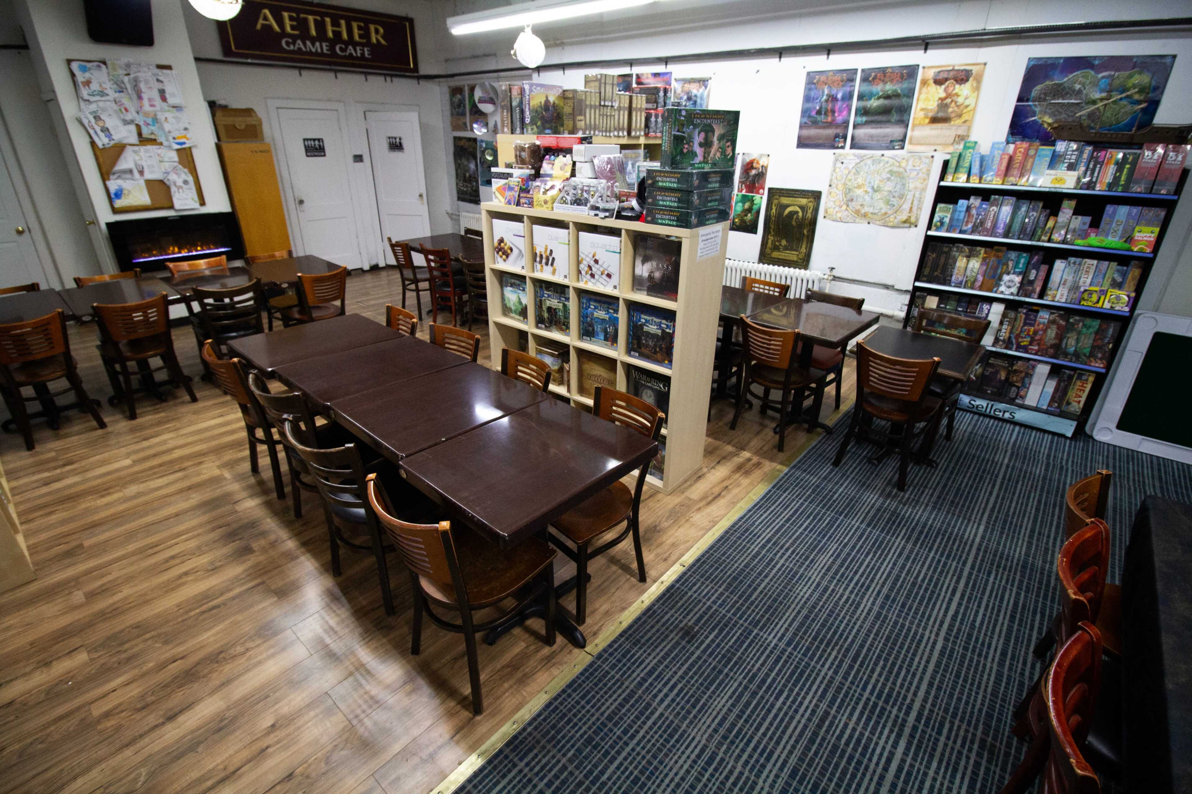 A game café with wooden tables, chairs, and shelves stocked with board games, along with a cozy seating area and posters on the walls.