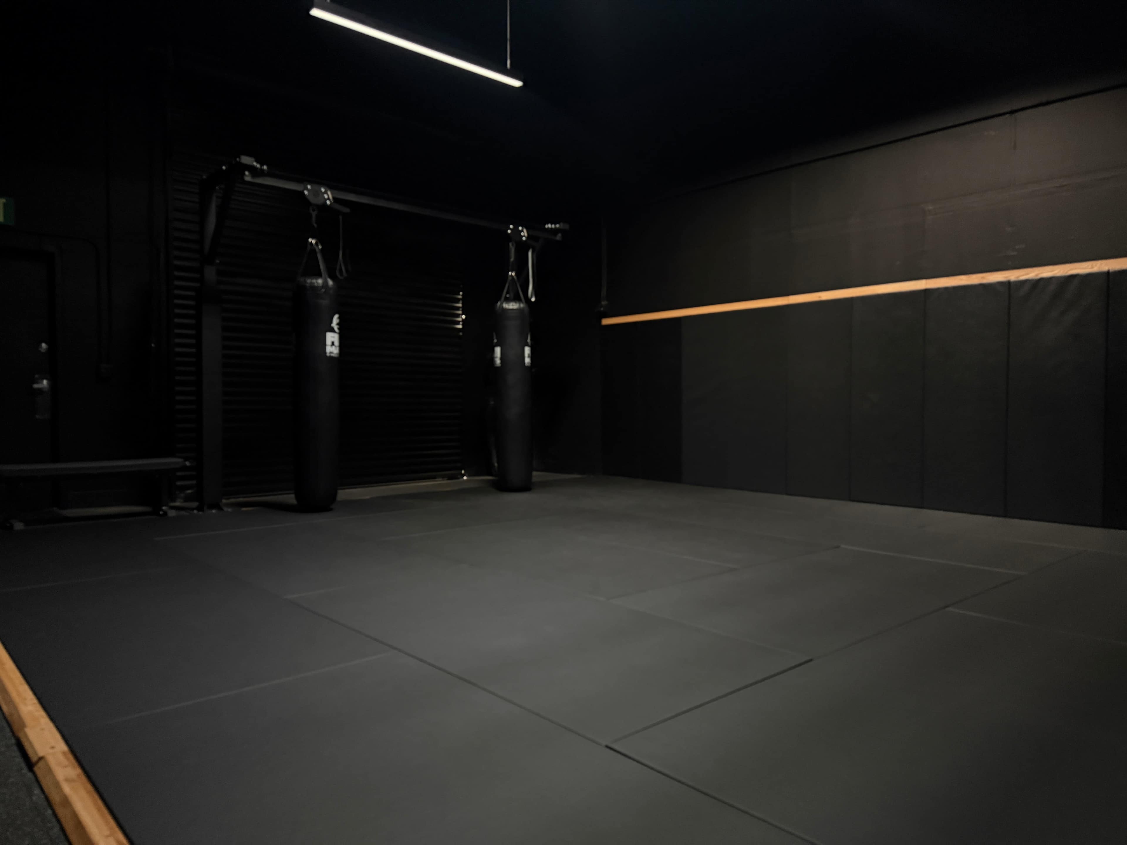 The image shows a sparsely furnished training room with black mats, two boxing bags suspended from the ceiling, and dark walls.