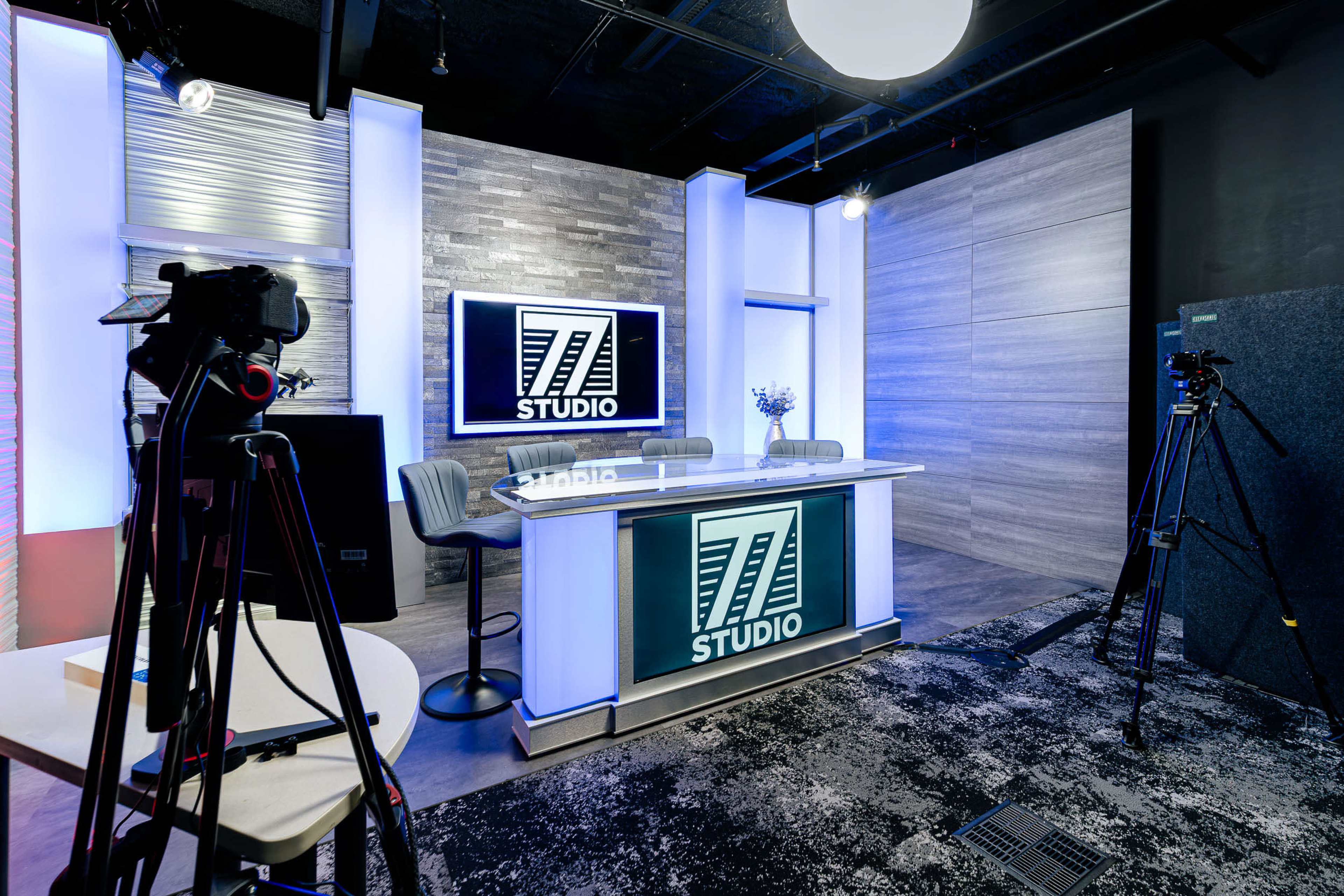 The image shows a modern studio setup with a broadcasting desk labeled "77 STUDIO," surrounded by cameras and a sleek wall design.
