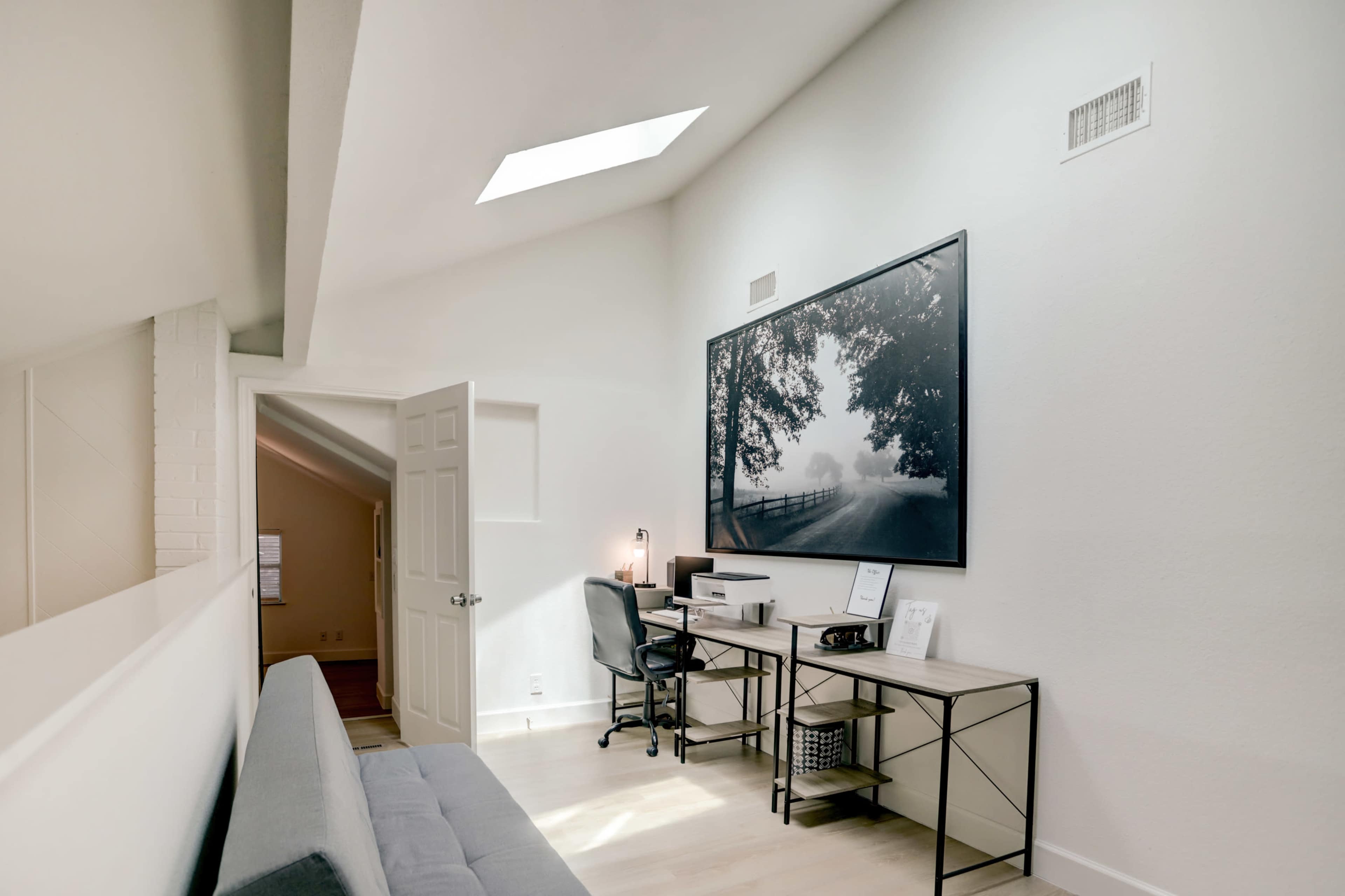 A bright room features a desk with a chair and a large black-and-white photograph on the wall, illuminated by natural light from a skylight.