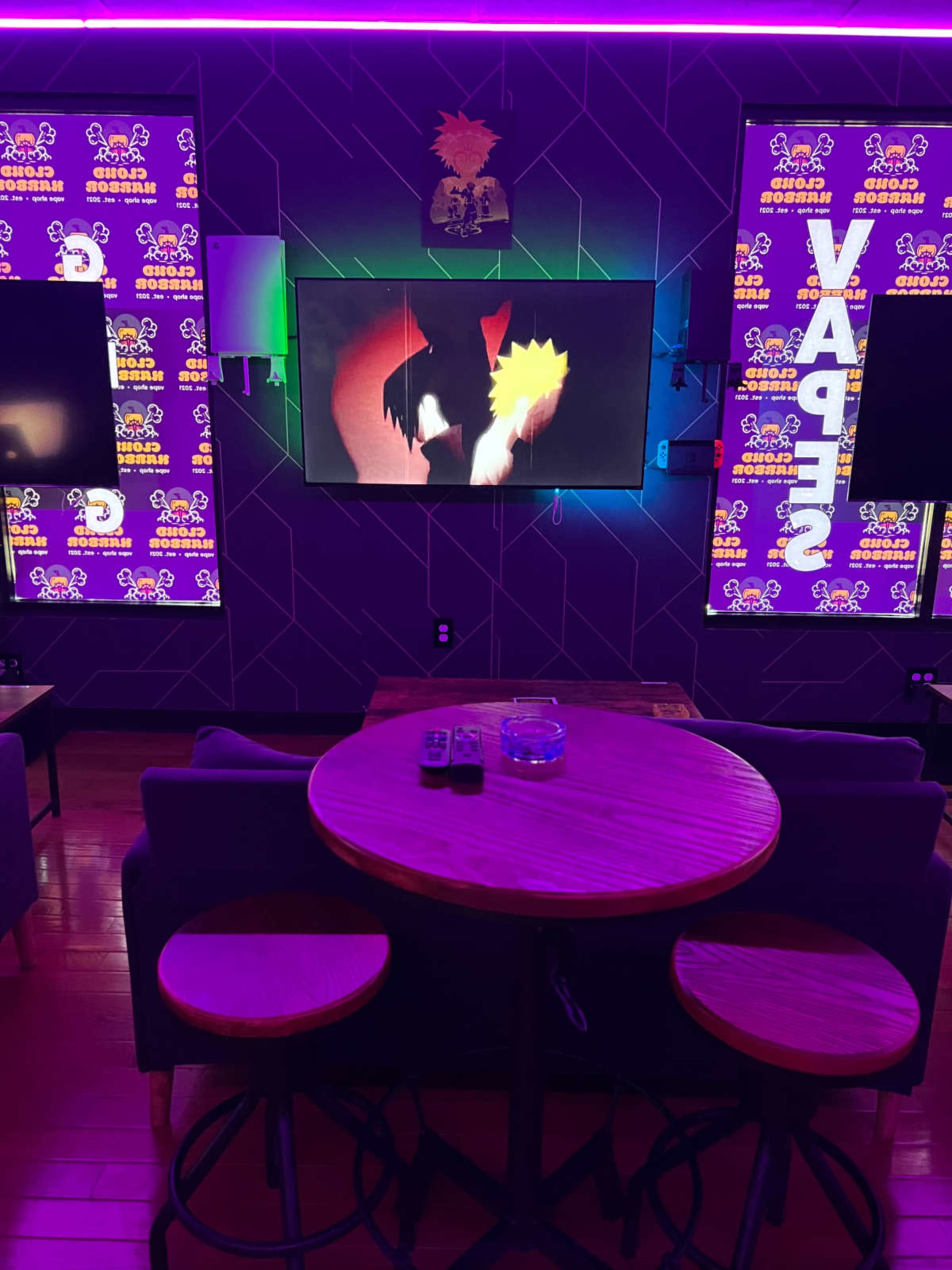 The image shows a modern entertainment room with a round wooden table, two stools, and multiple screens displaying colorful wall art and a TV screen featuring animated content.