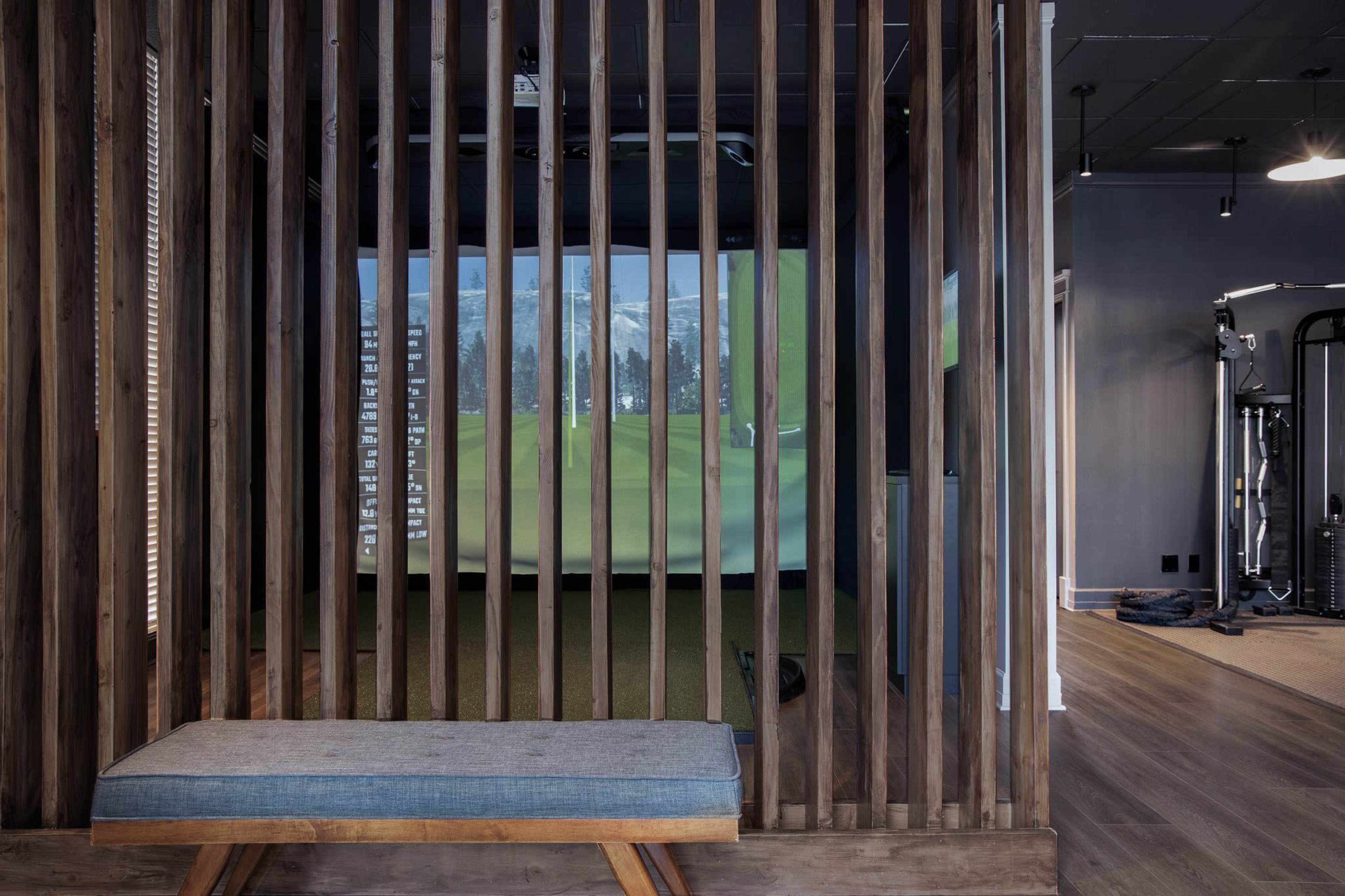A modern indoor golf simulation area separated by wooden slats from an adjacent gym space.