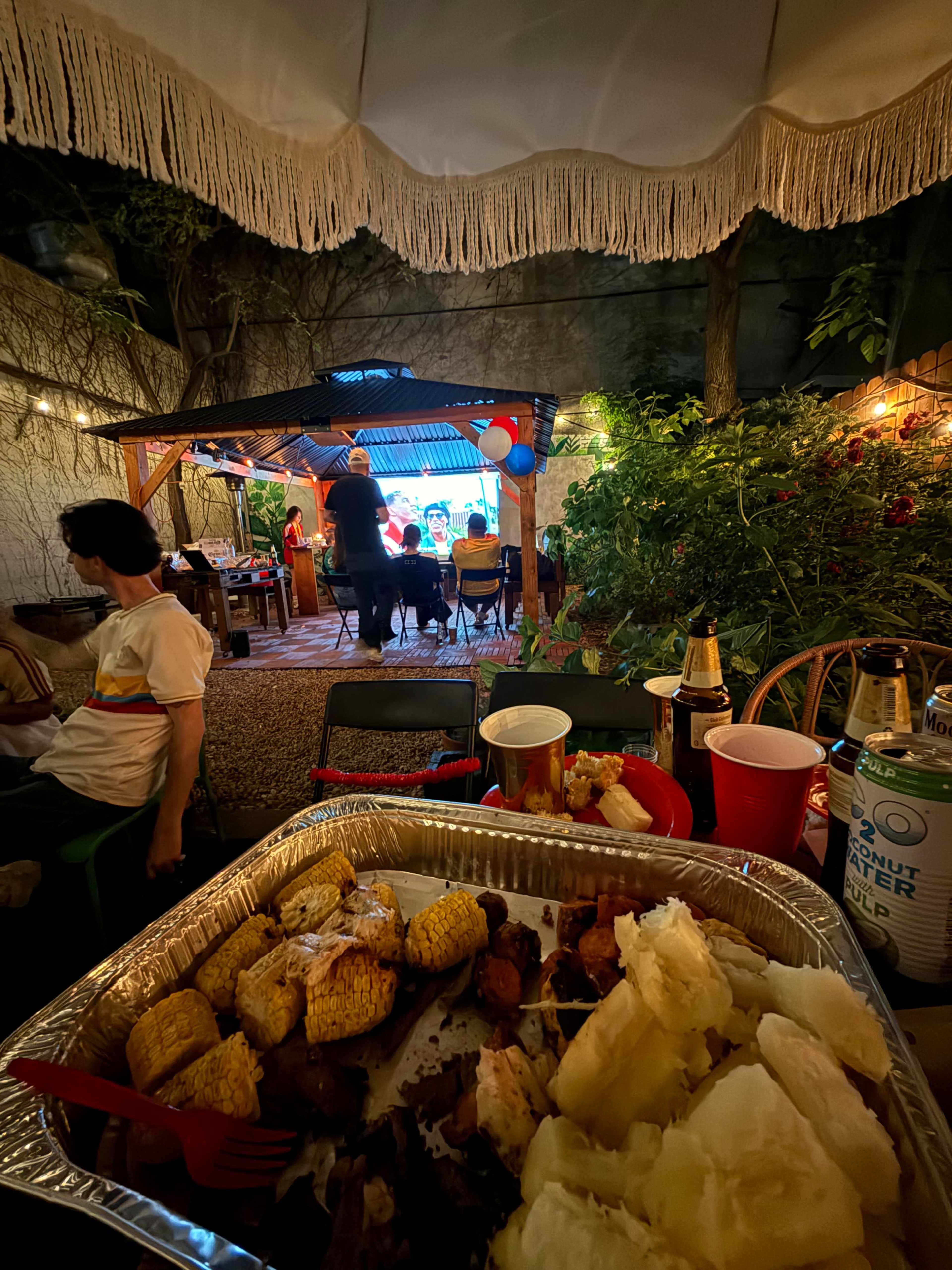 A backyard gathering takes place under a canopy, featuring people seated at a table with food, while a projector screen displays a scene in the background.