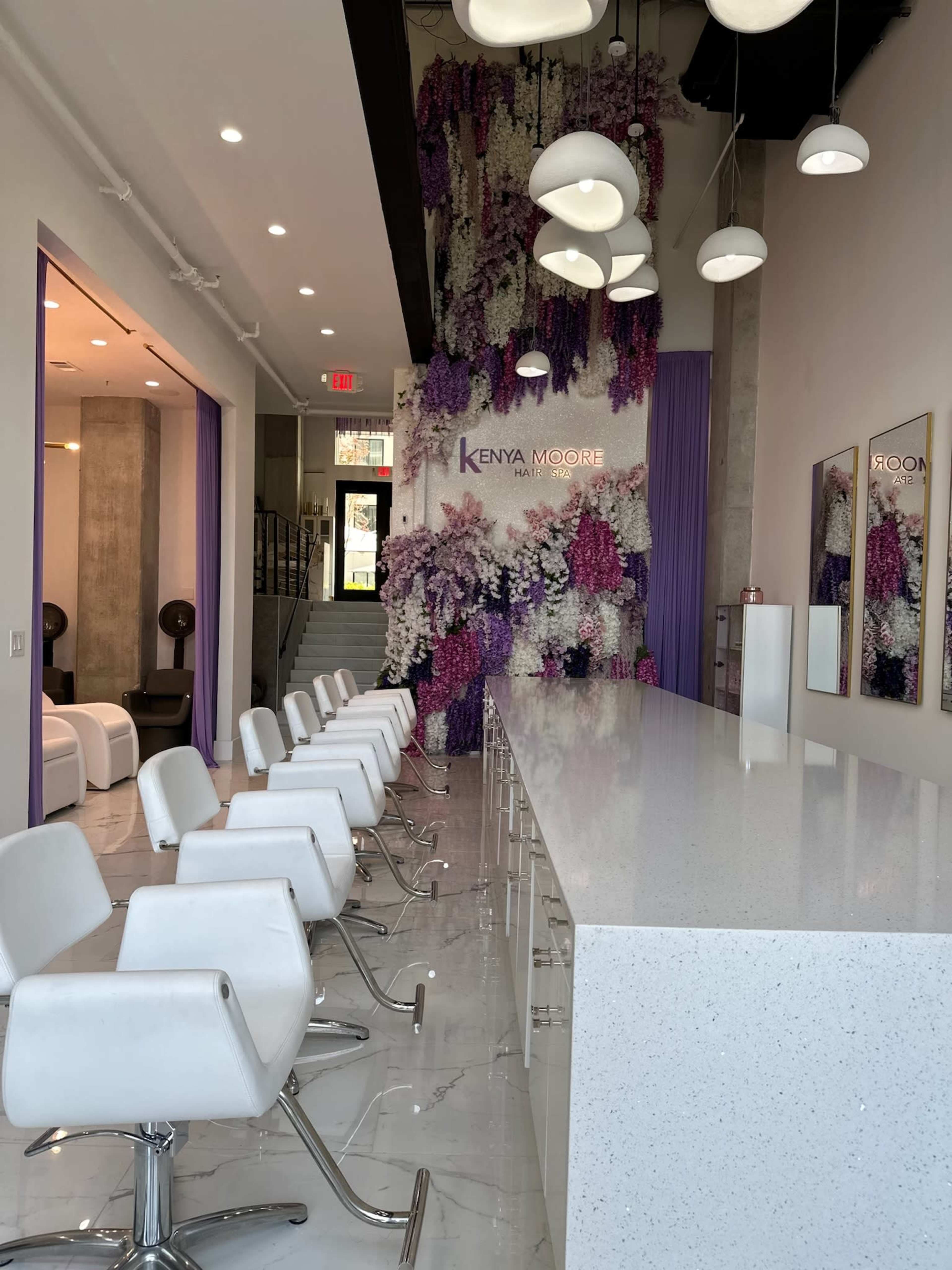A modern salon features a sleek white countertop with white bar stools, surrounded by a wall adorned with purple and white floral arrangements.
