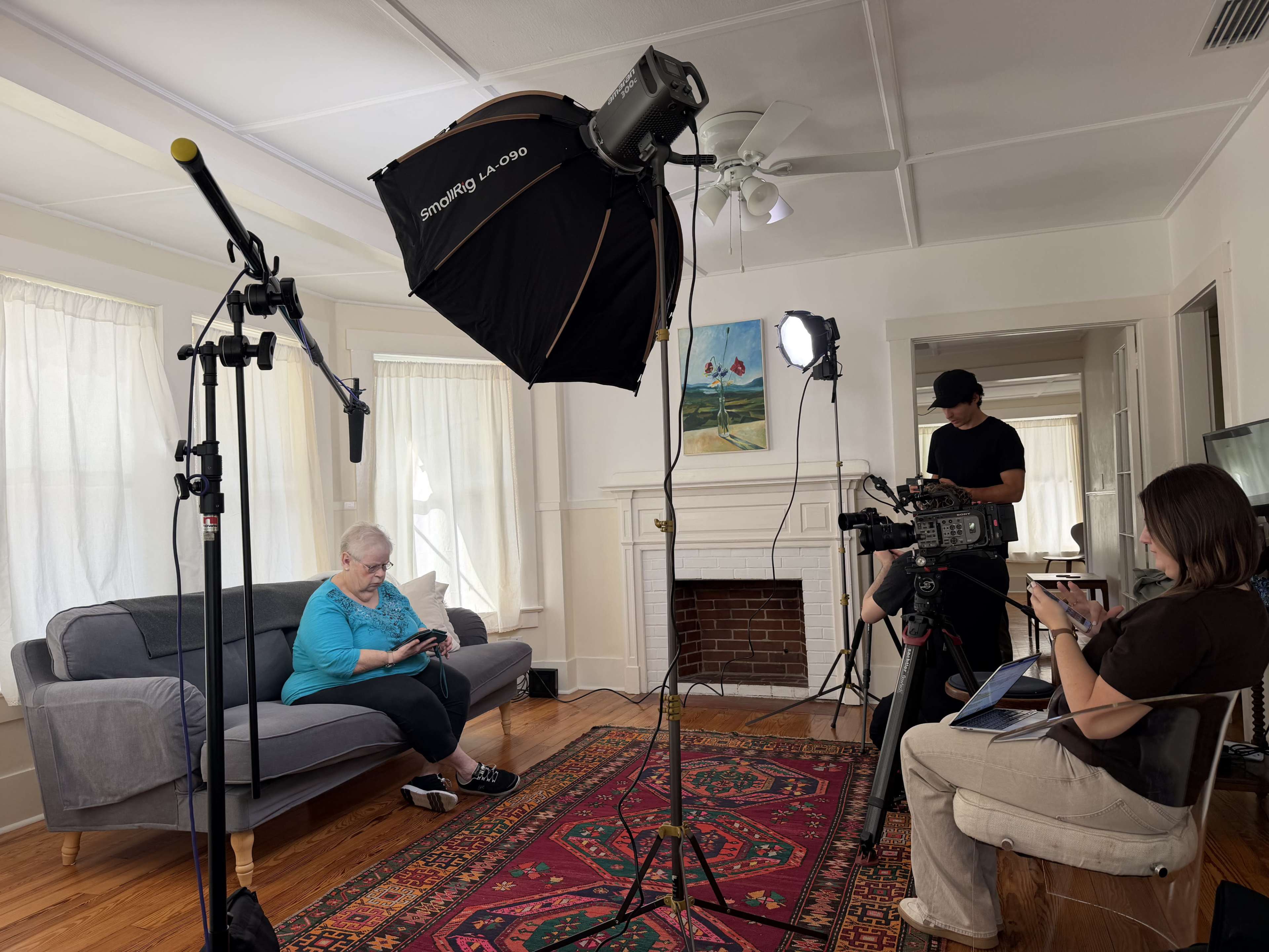 A woman sits on a gray couch in a well-lit living room, while two crew members set up filming equipment nearby.