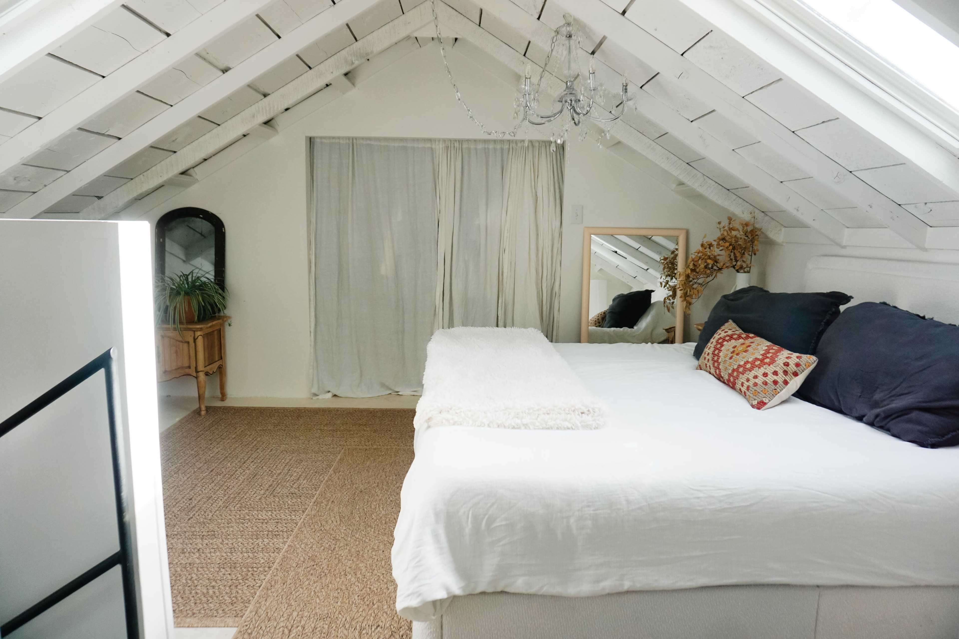The image shows a cozy attic bedroom with a sloped ceiling, a large bed adorned with pillows, and a mirror reflecting natural light from a window.