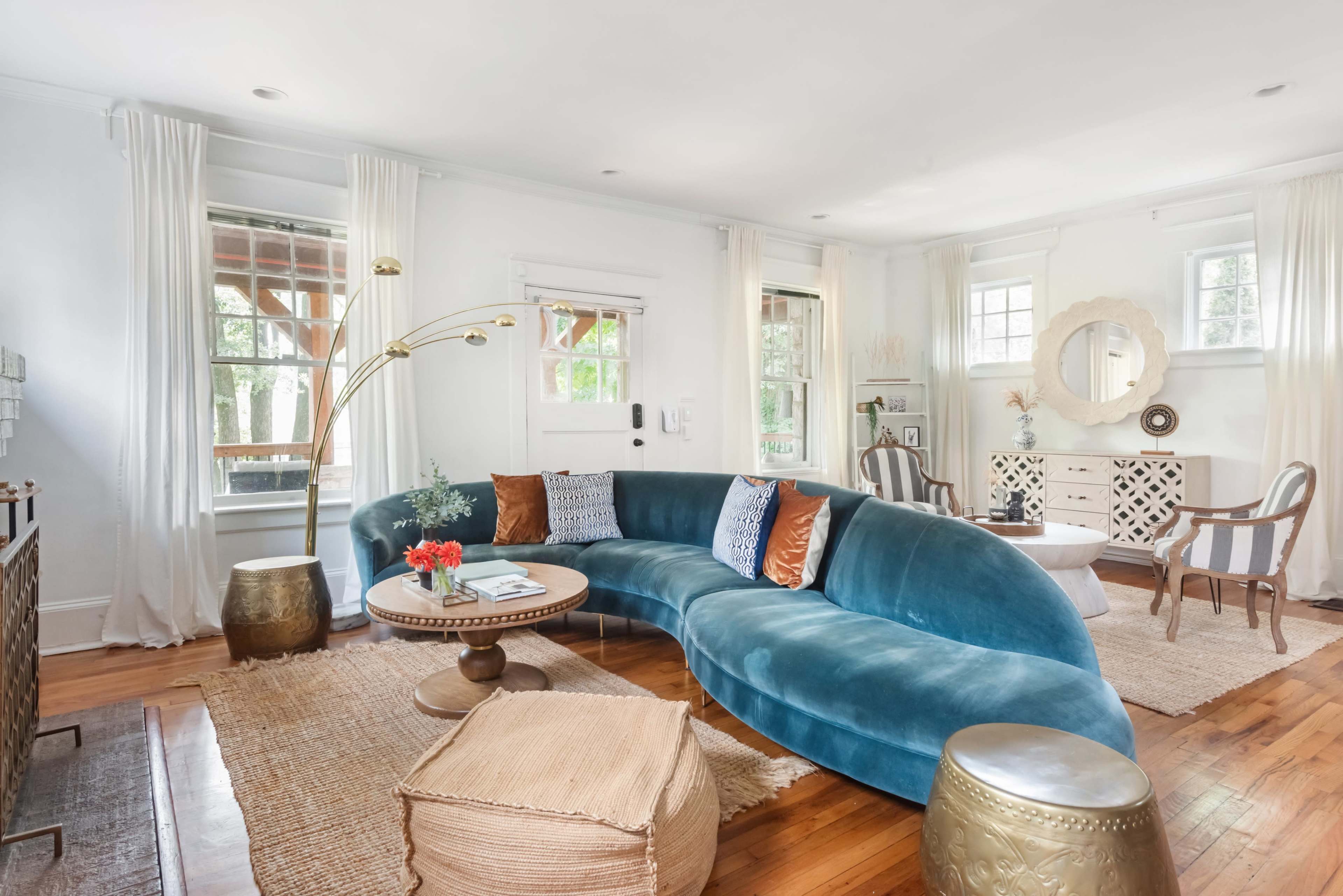 The living room features a curved, blue velvet sofa, a round coffee table, decorative pillows, and large windows letting in natural light, complemented by a woven rug and brass side tables.