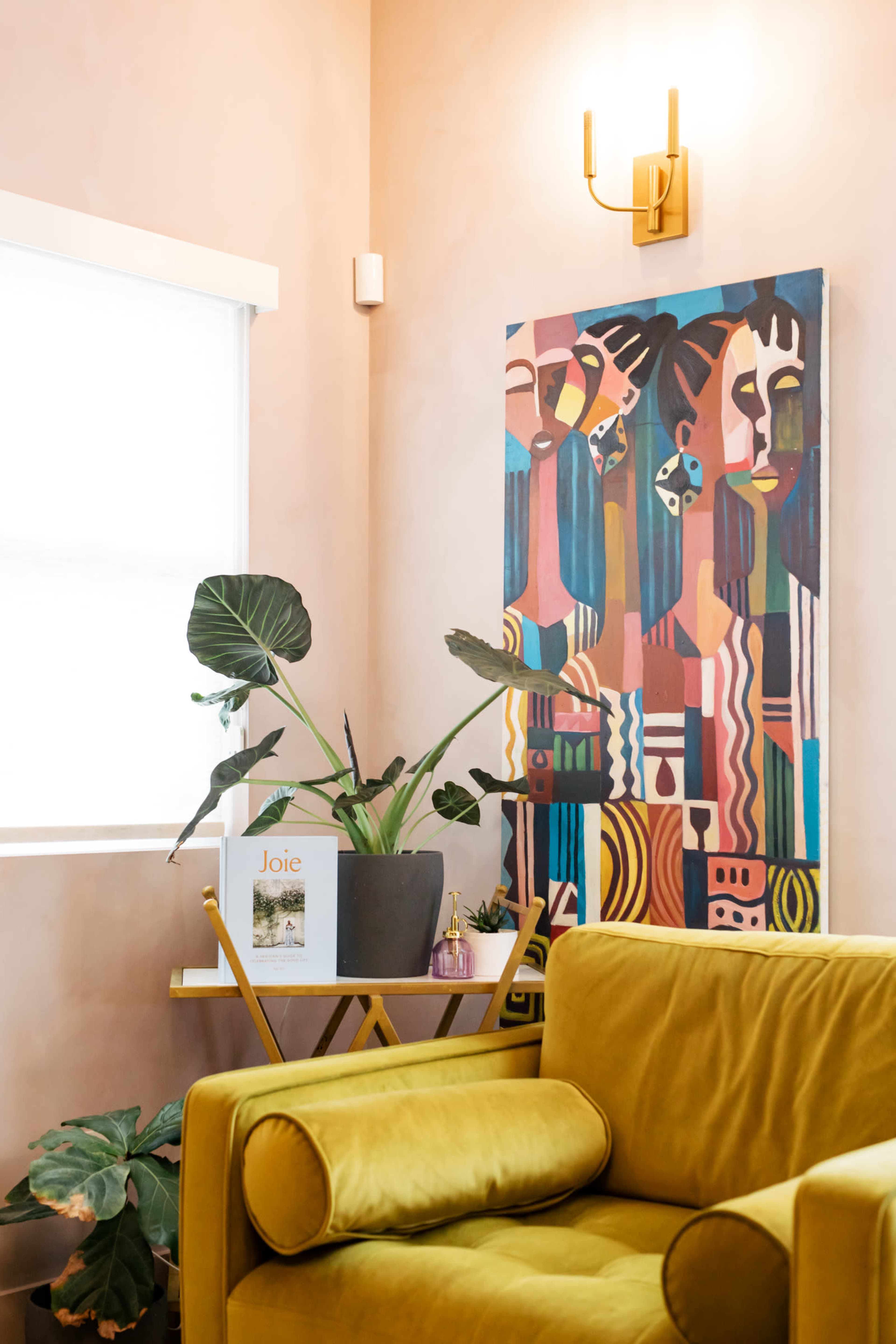 The image shows a cozy corner of a room featuring a yellow couch, a small table with a book, a potted plant, and a colorful abstract painting on the wall.