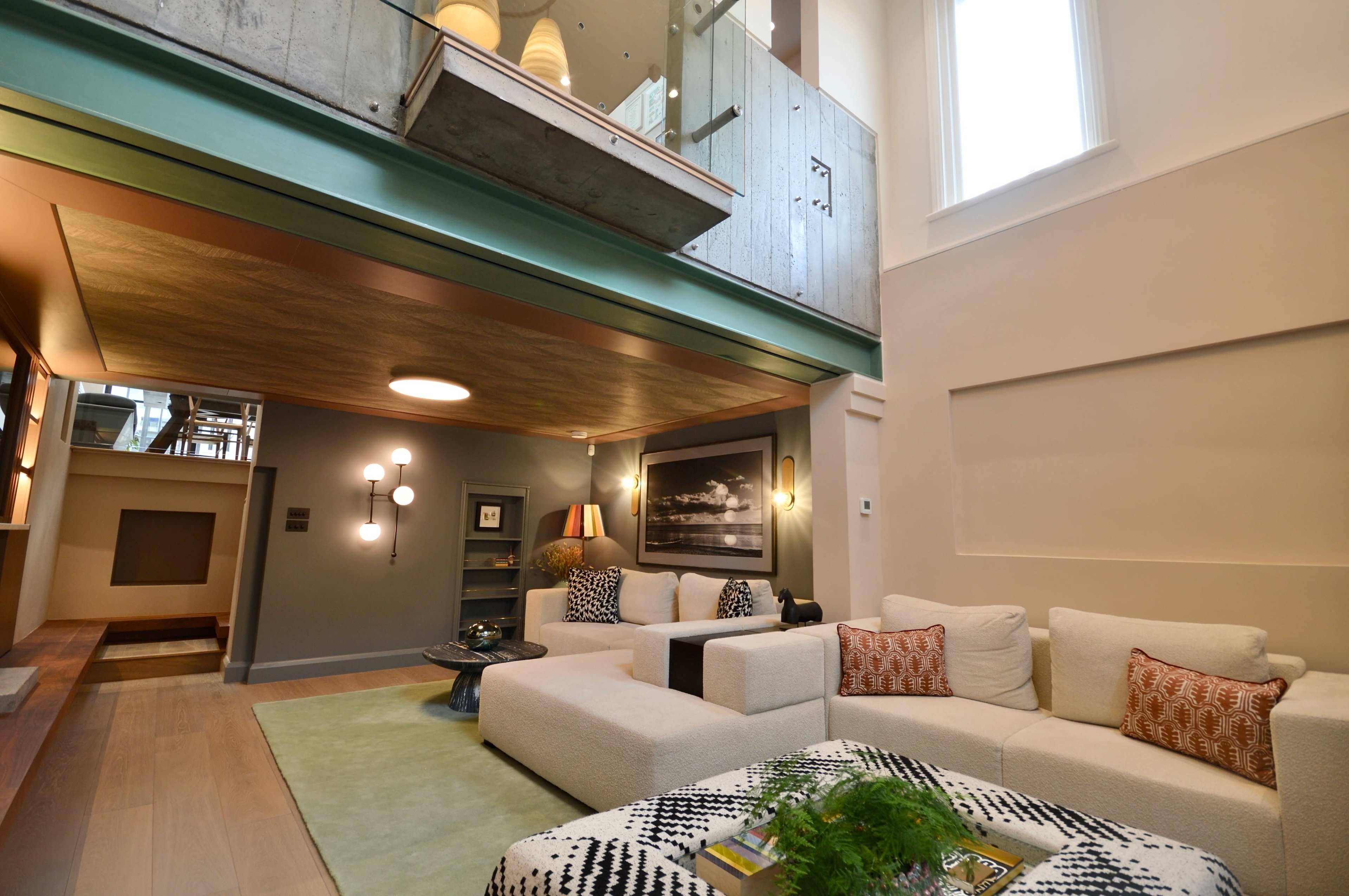 The image shows a modern living room with a large sectional sofa, a coffee table, and an open layout that leads to a staircase and a second-level balcony.