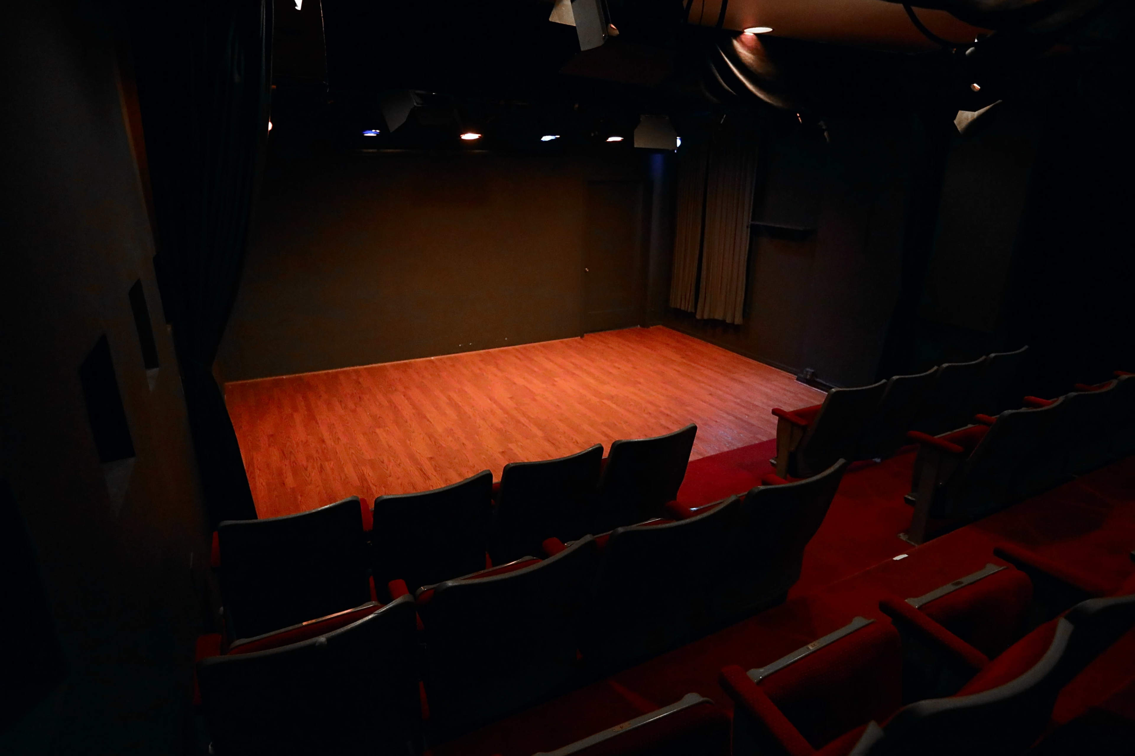 The image shows a dimly lit theater with a wooden stage and empty red seats facing it.