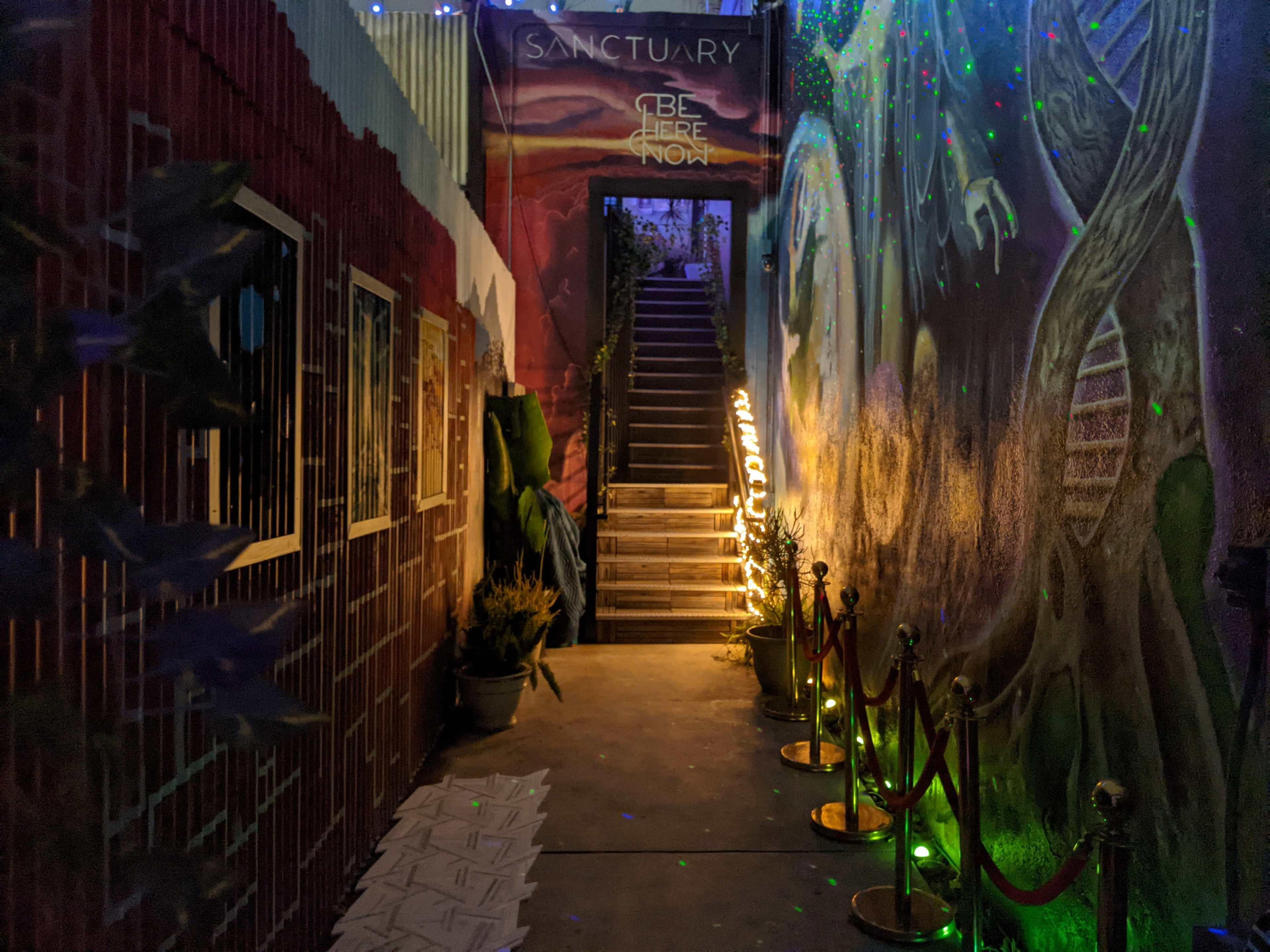 The image shows a dimly lit hallway with a staircase at the end, flanked by murals and decorative lights, and lined with velvet ropes.