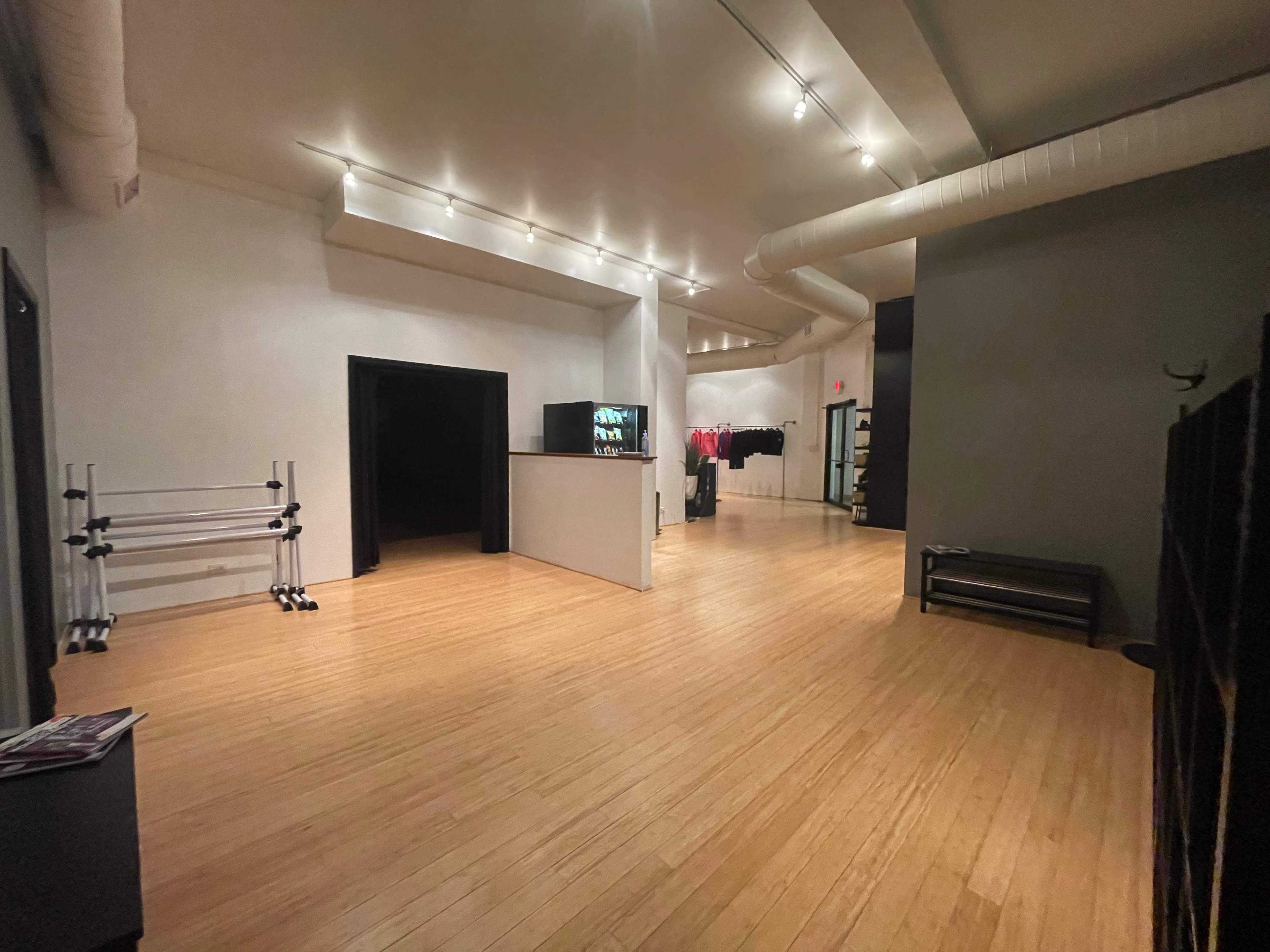 The image shows a spacious, empty room with wooden flooring and minimalist decor, featuring a reception area and clothing racks along the walls.