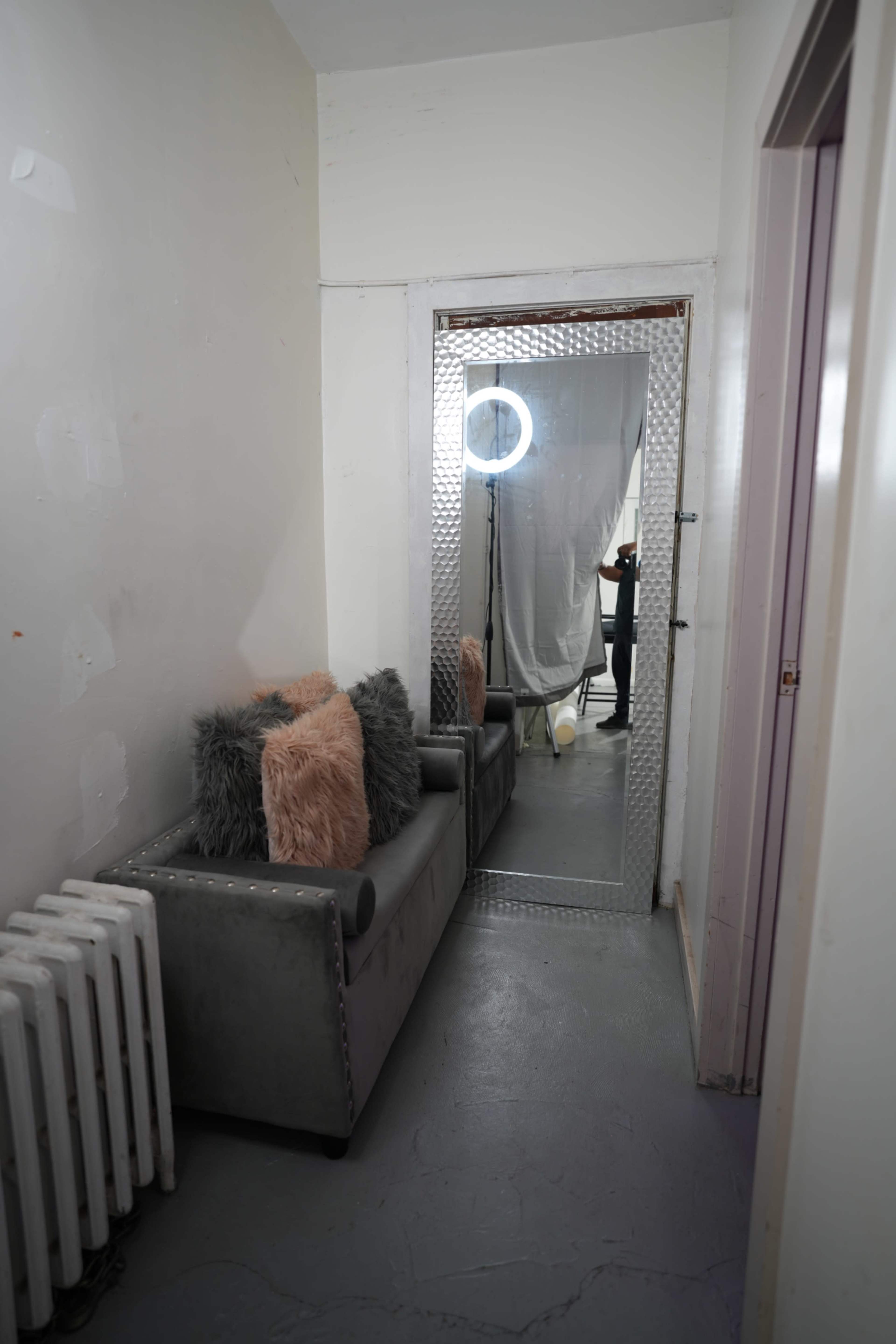 A narrow hallway features a gray couch with decorative pillows, a radiator on the left, and a mirror with a ring light at the end of the corridor.