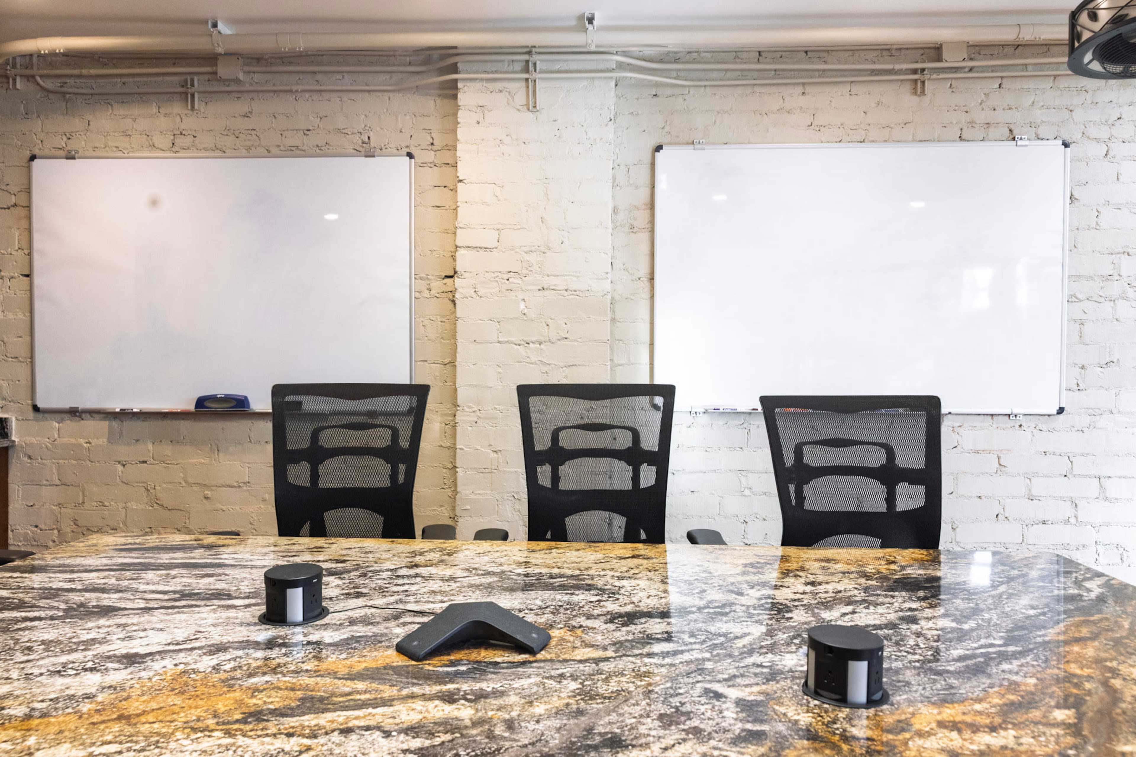 A conference room features a large granite table with three black office chairs, flanked by two whiteboards on the wall.