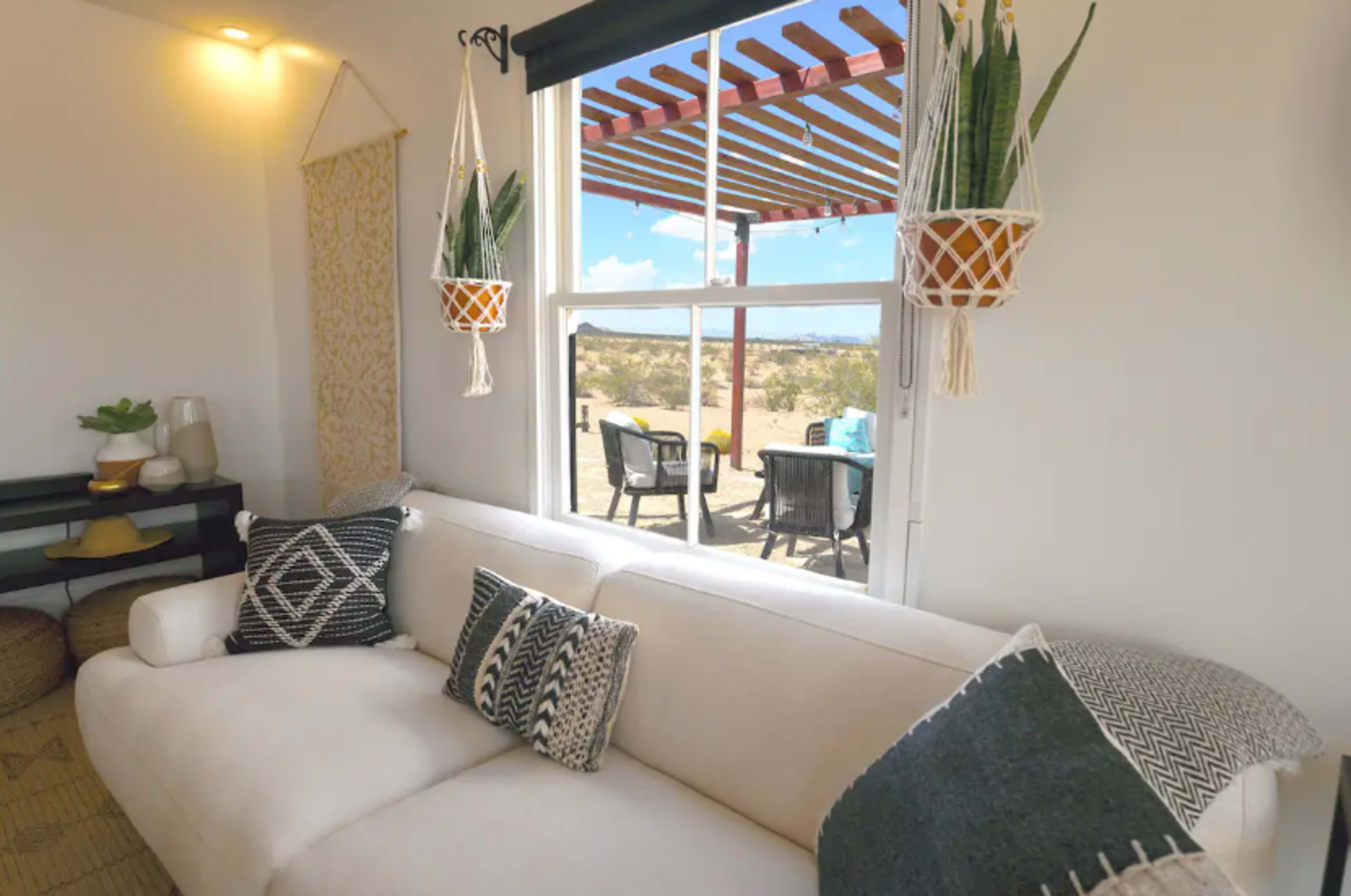 A light-filled room features a white sofa, potted plants, and hanging planters, with a view of an outdoor seating area through a large window.