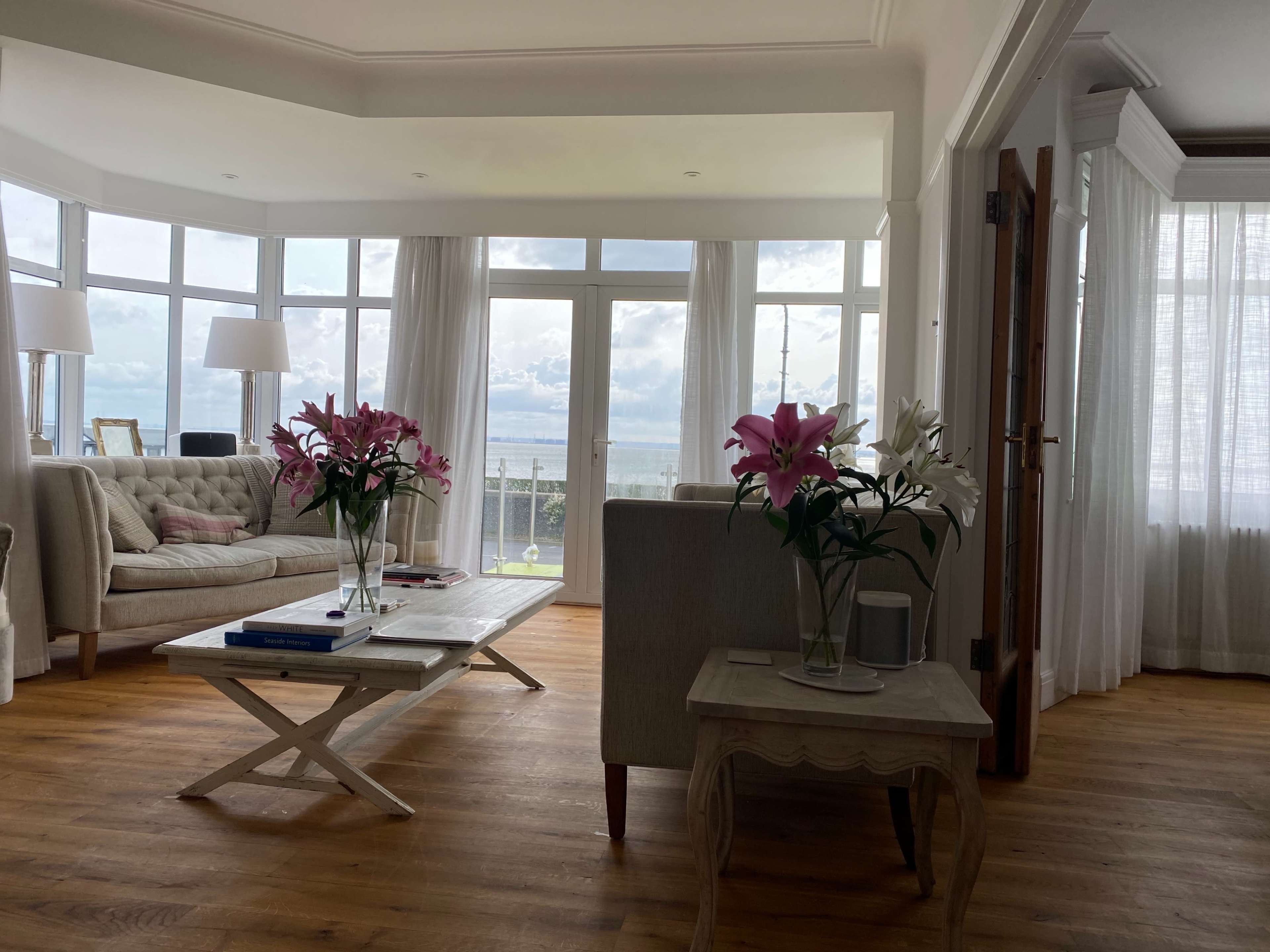 A bright living room features a beige sofa, a coffee table with books, and large windows offering a view of the ocean.