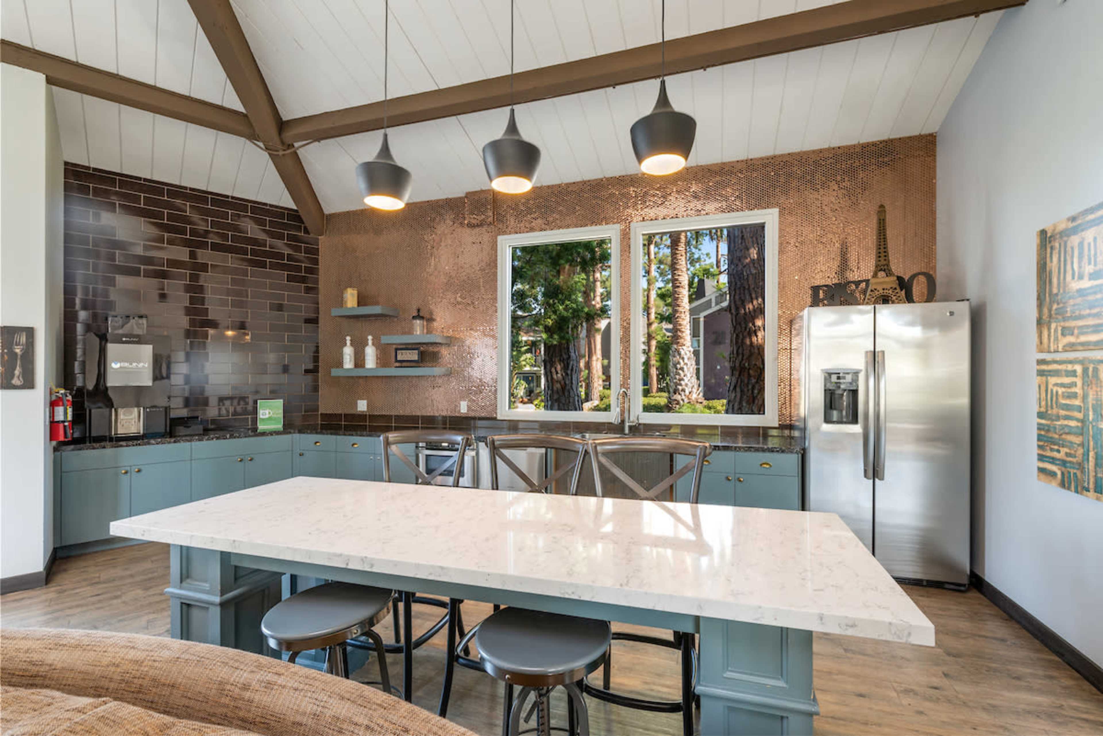 A modern kitchen features a large white countertop island, copper backsplash, and stainless steel refrigerator, with large windows showcasing greenery outside.