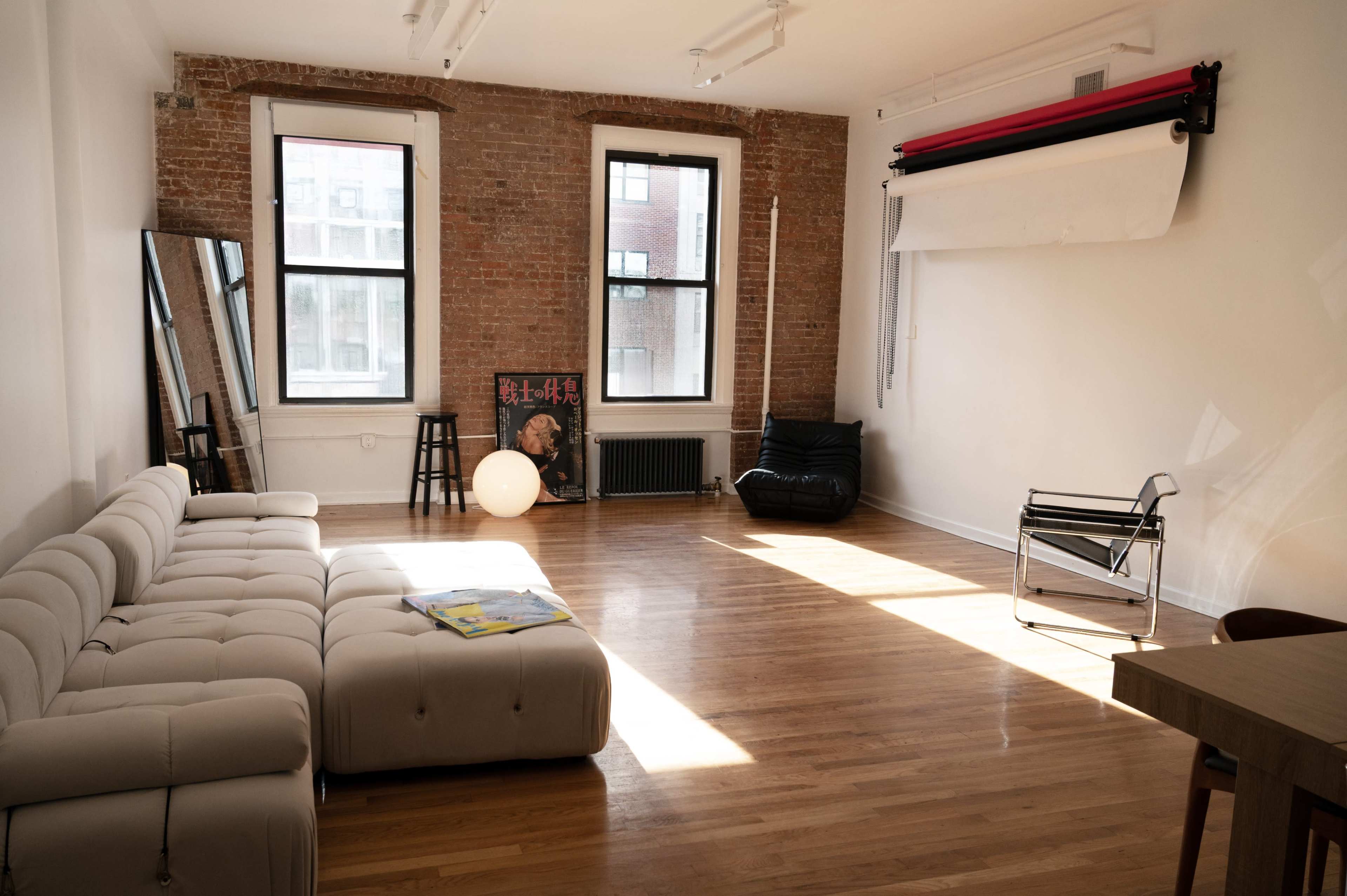 The image shows a spacious, well-lit room with hardwood floors, featuring a white leather sectional sofa, a black lounge chair, and large windows revealing a brick wall.