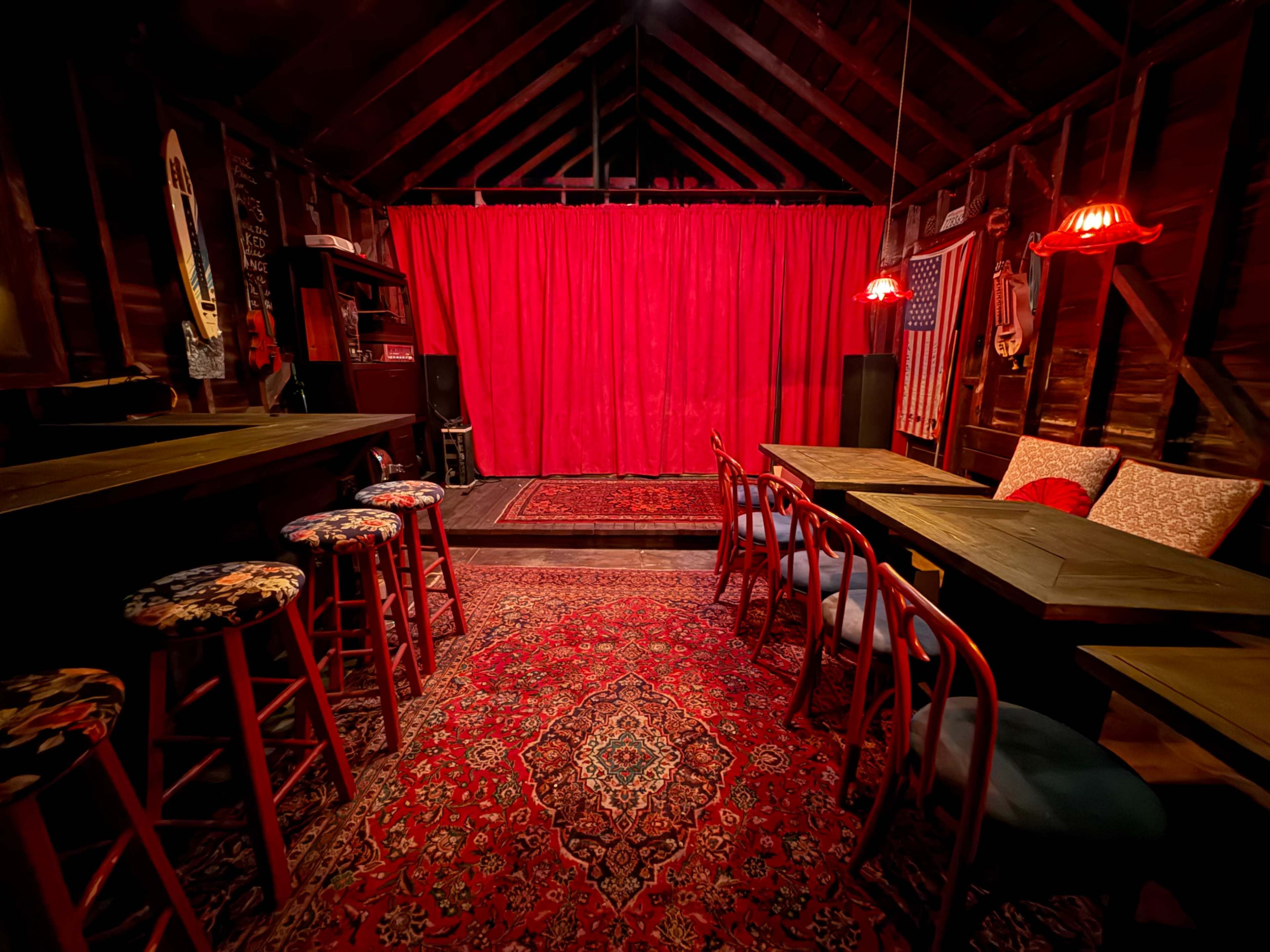 The image shows an intimate performance space with a red curtain at the front, wooden tables, and stools arranged on a patterned rug surrounded by rustic wood walls.