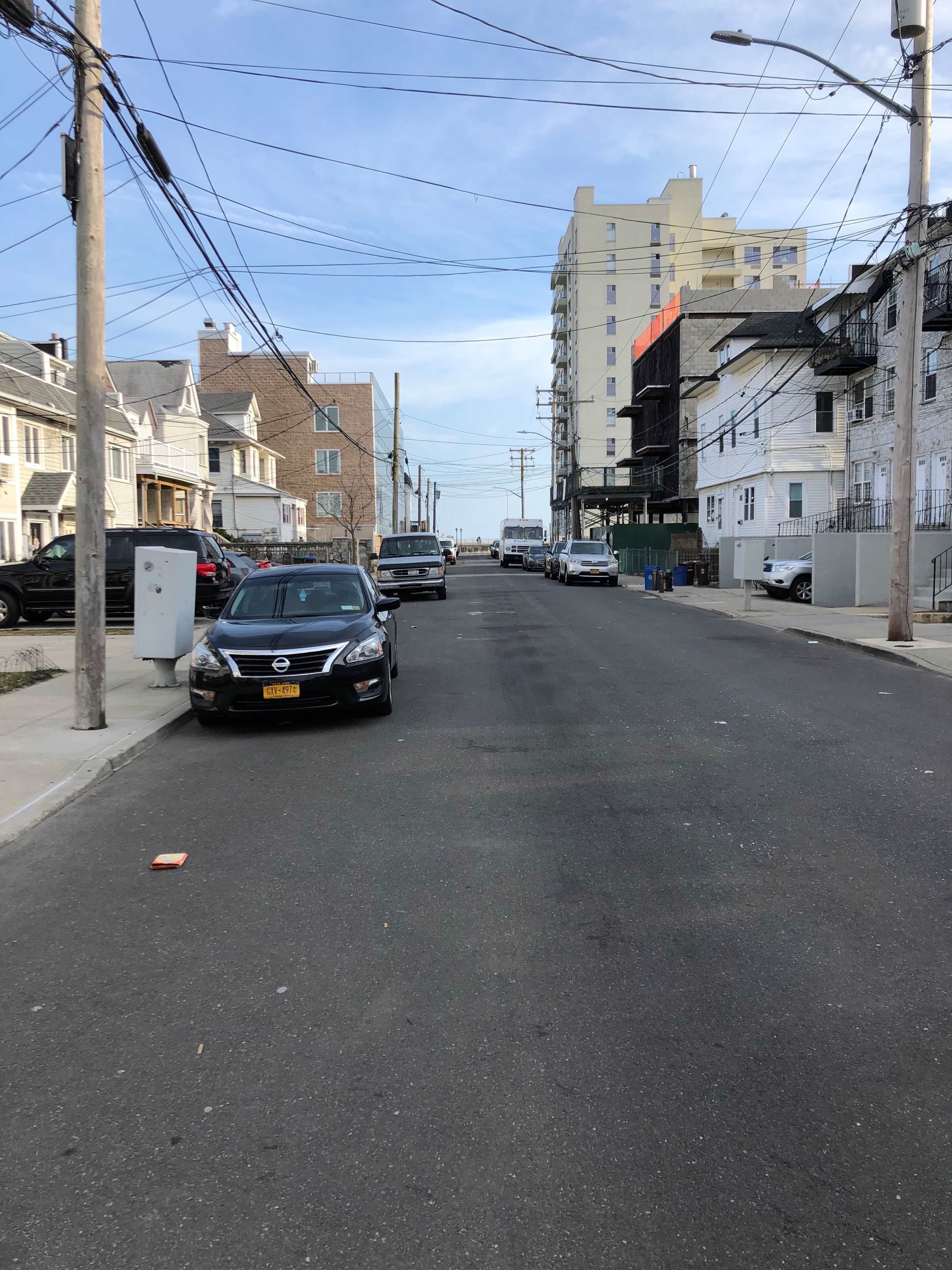 A wide, empty street lined with parked cars and buildings, with power lines stretching overhead under a clear blue sky.