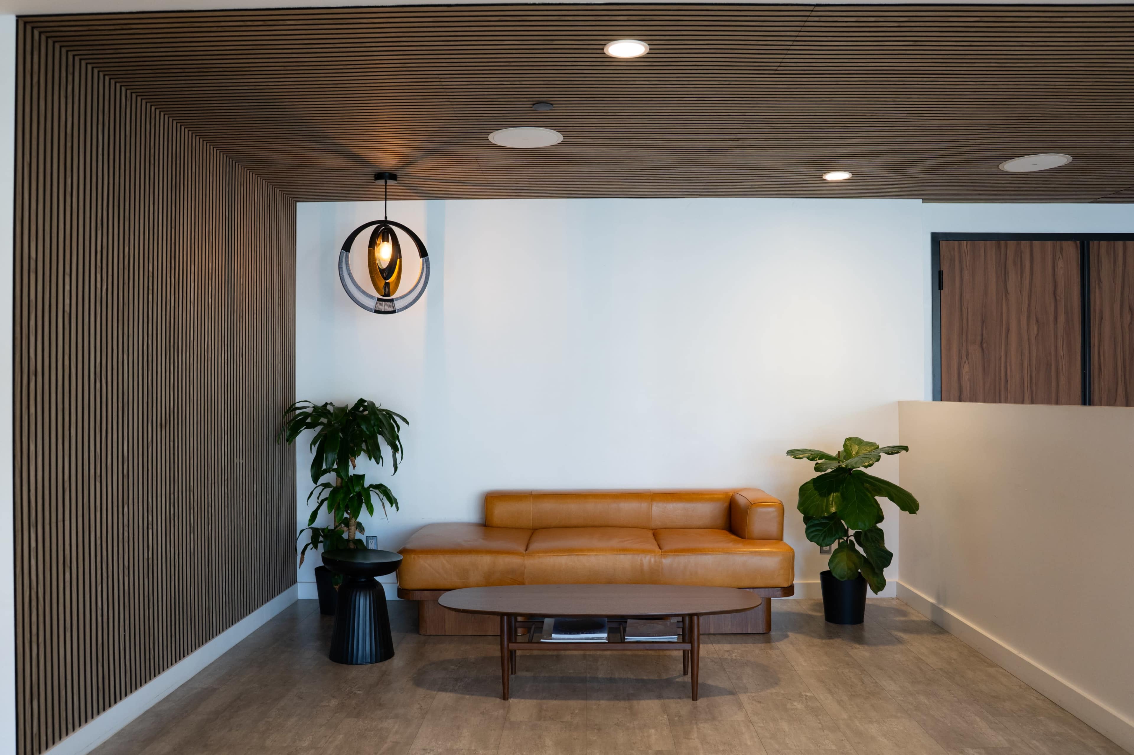 The image shows a modern interior space featuring a brown leather sofa, a round coffee table, and two decorative plants against a backdrop of wood paneling and neutral walls.