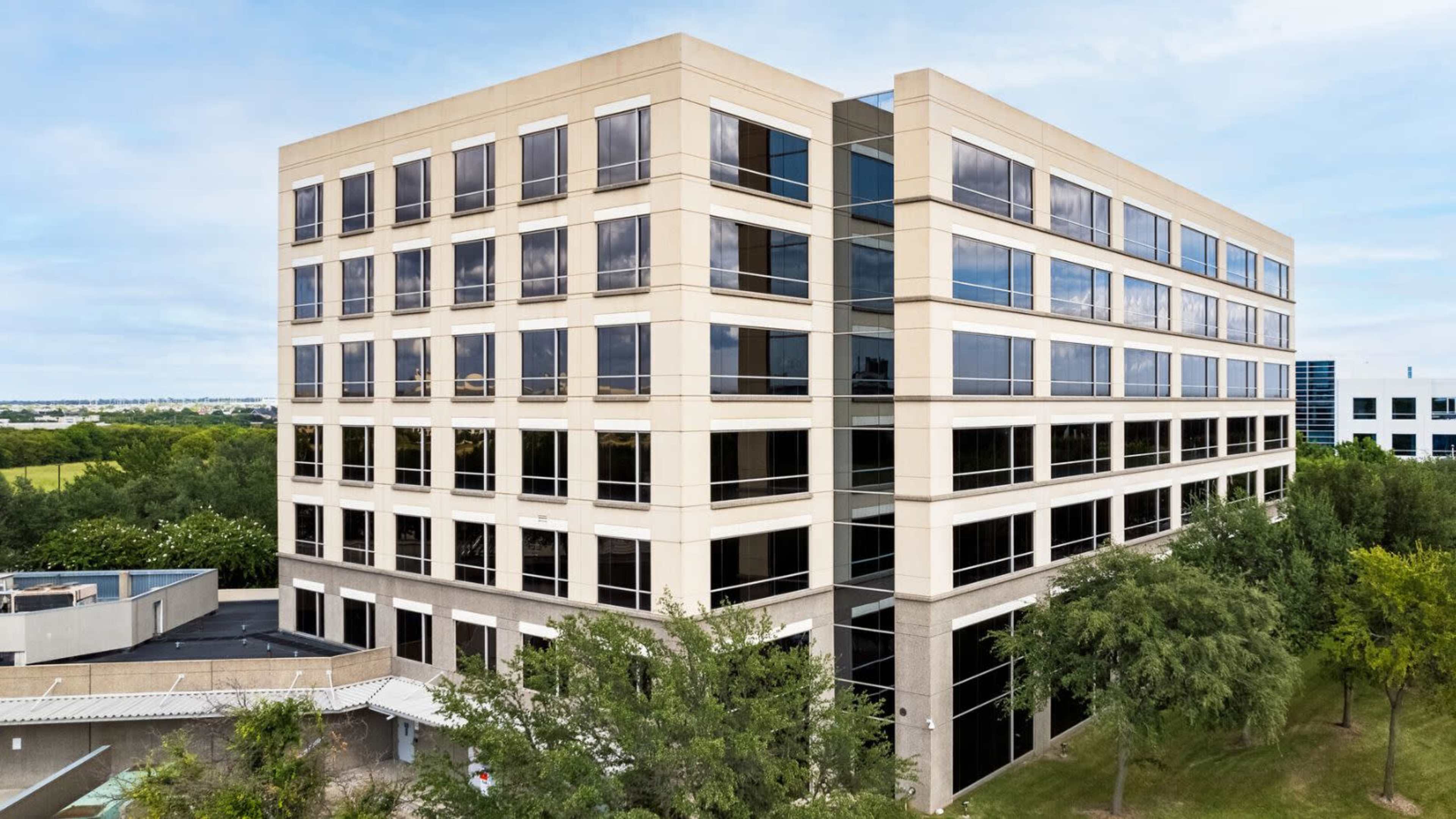 The image shows a multi-story office building with large windows and a landscaped area featuring trees.