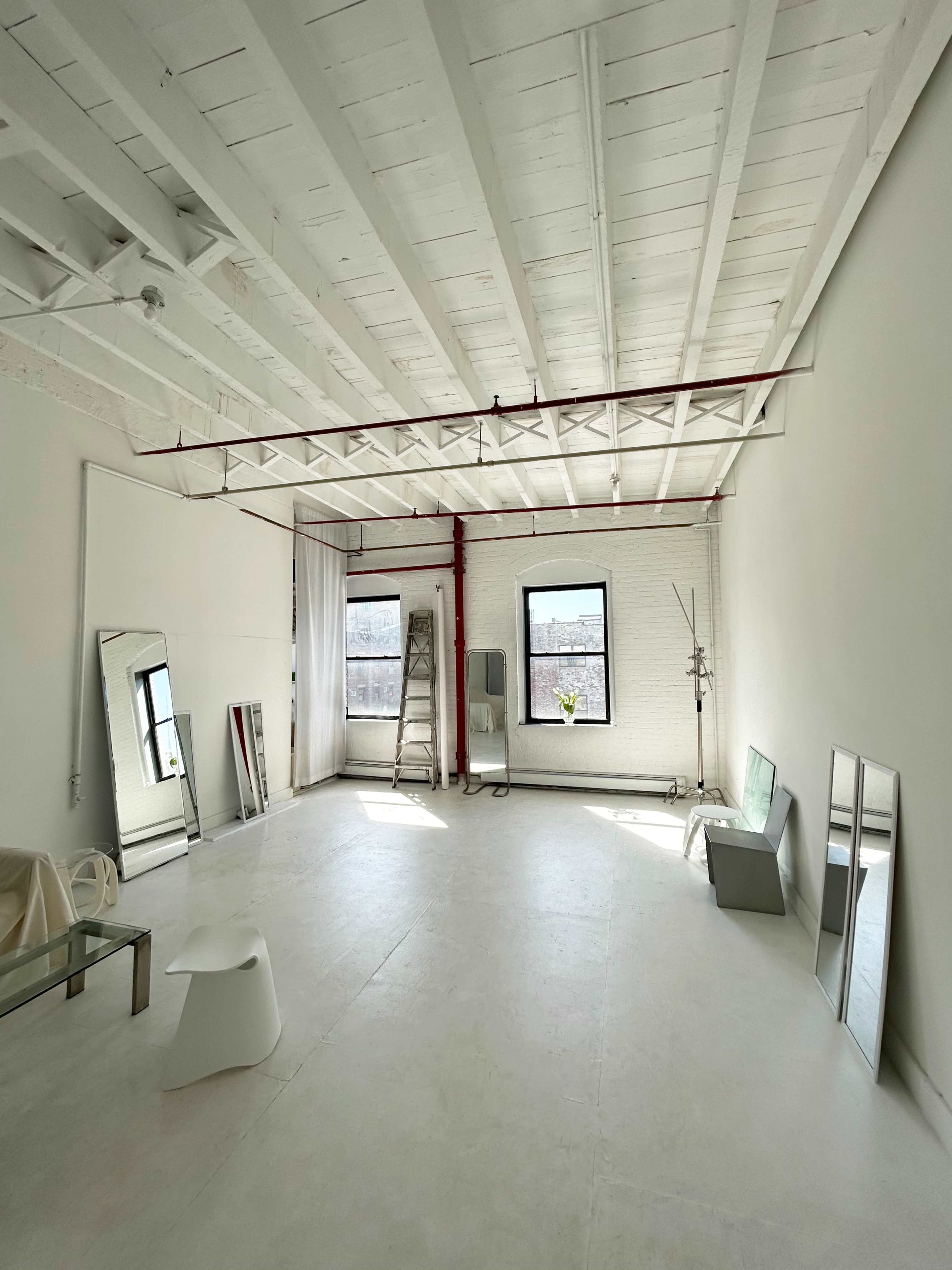 A spacious, minimalist studio with white walls, wooden beams, two large mirrors, a folding ladder, and large windows providing natural light.