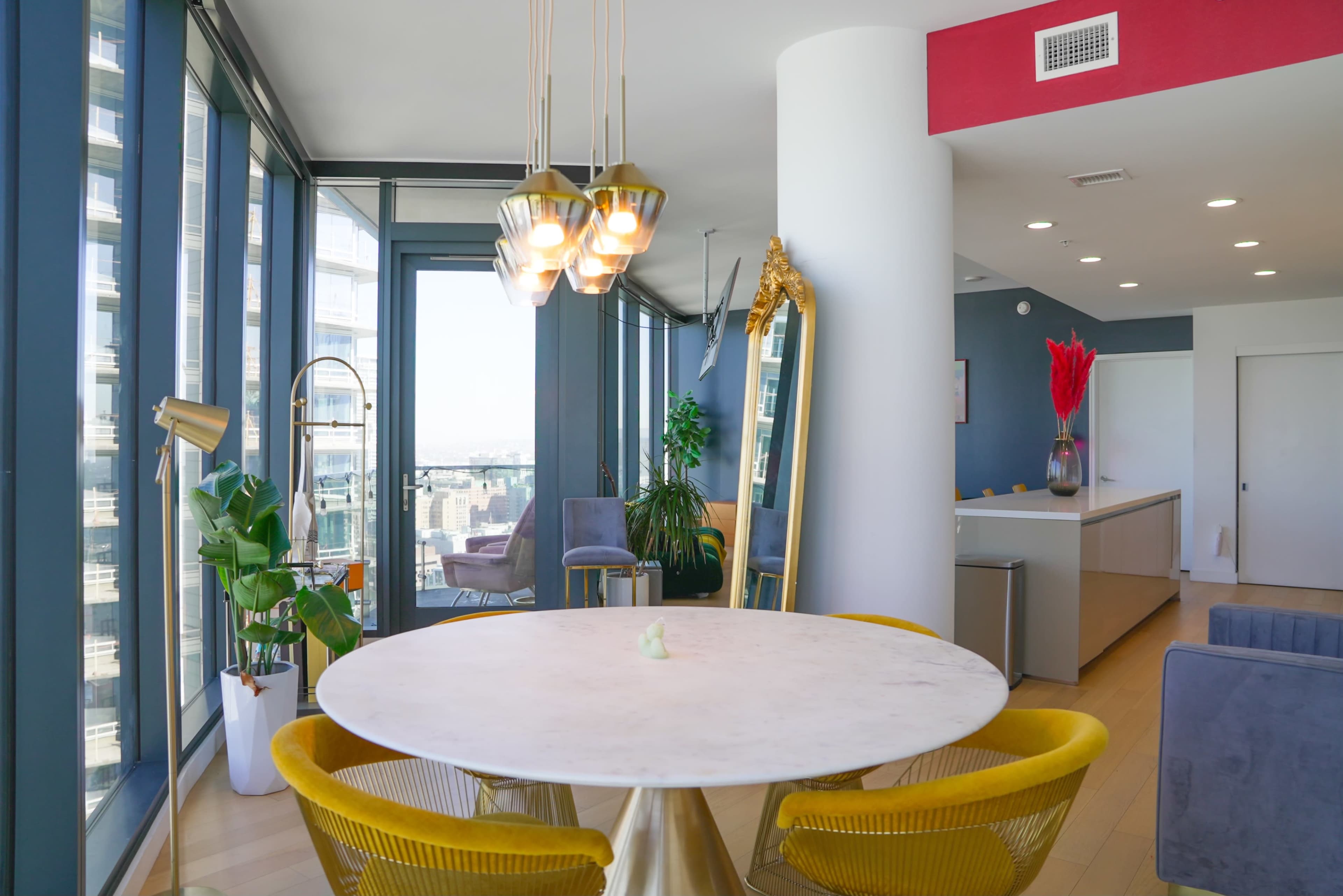 A modern dining area features a round marble table surrounded by yellow upholstered chairs, with large windows offering a view of the cityscape.
