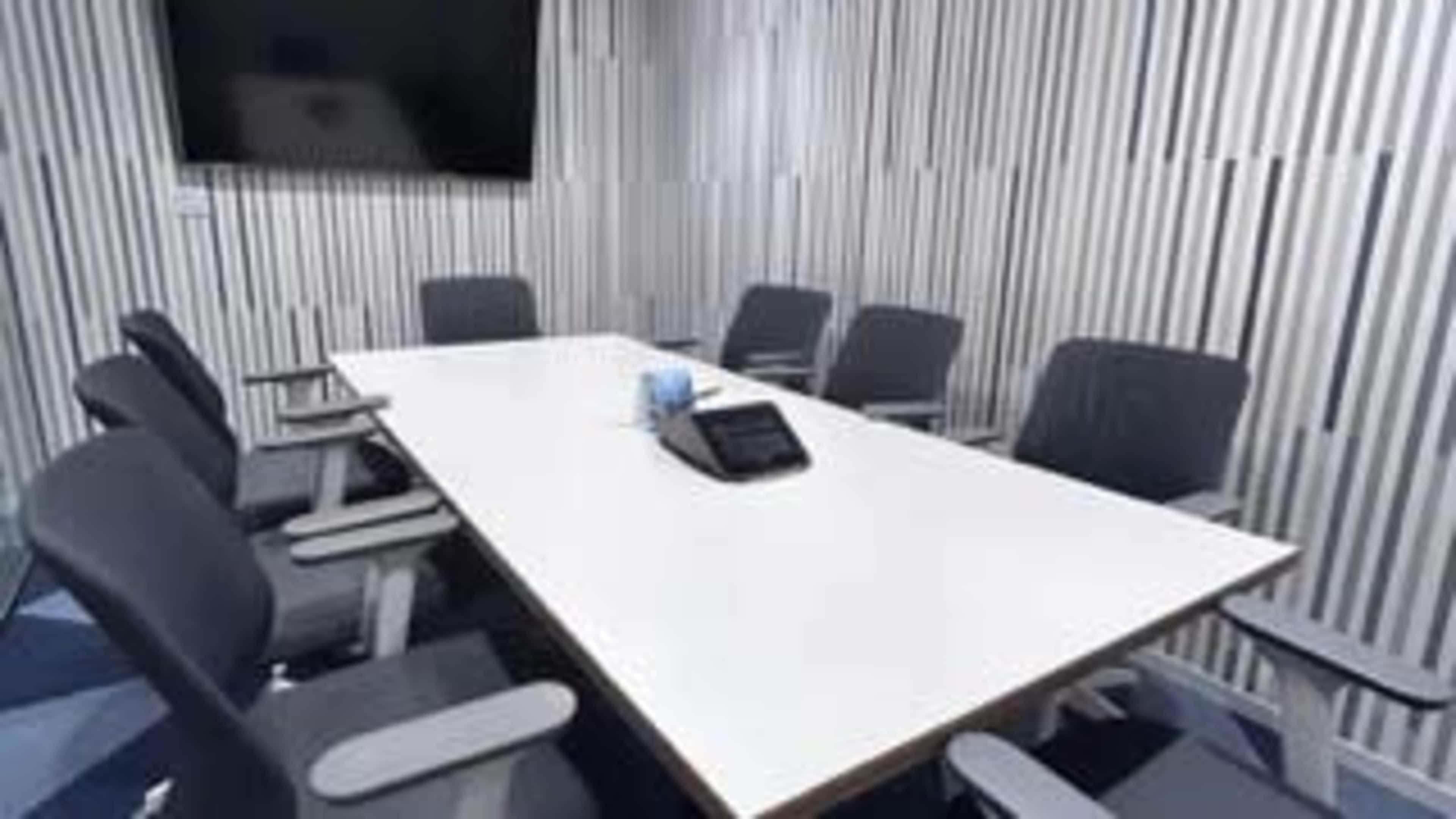 A modern conference room features a long white table surrounded by gray chairs, with a television mounted on the wall and a conference phone placed at the center.