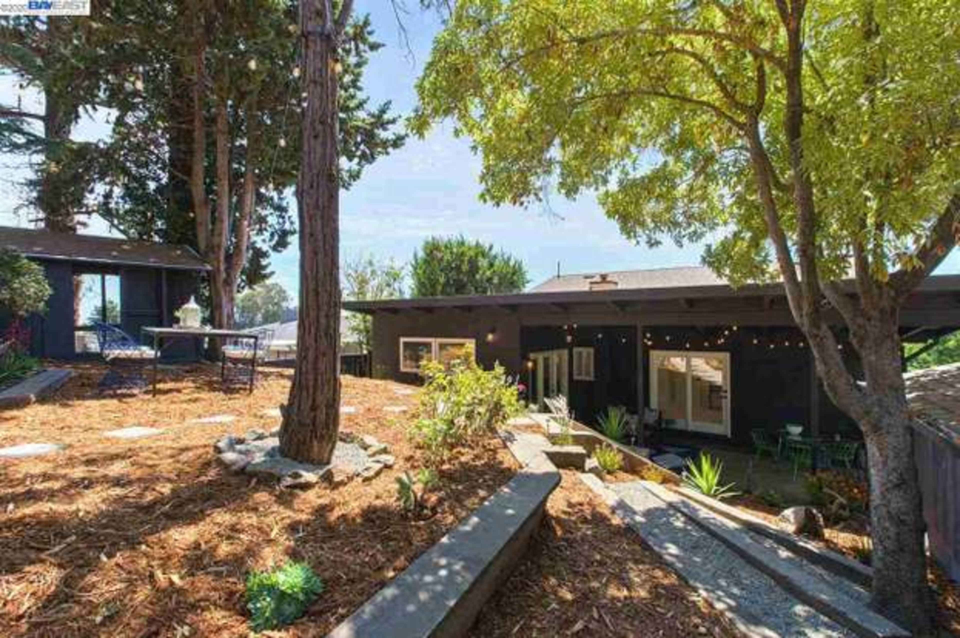 A modern single-story house surrounded by trees and landscaped grounds, with a gravel pathway leading to the entrance.