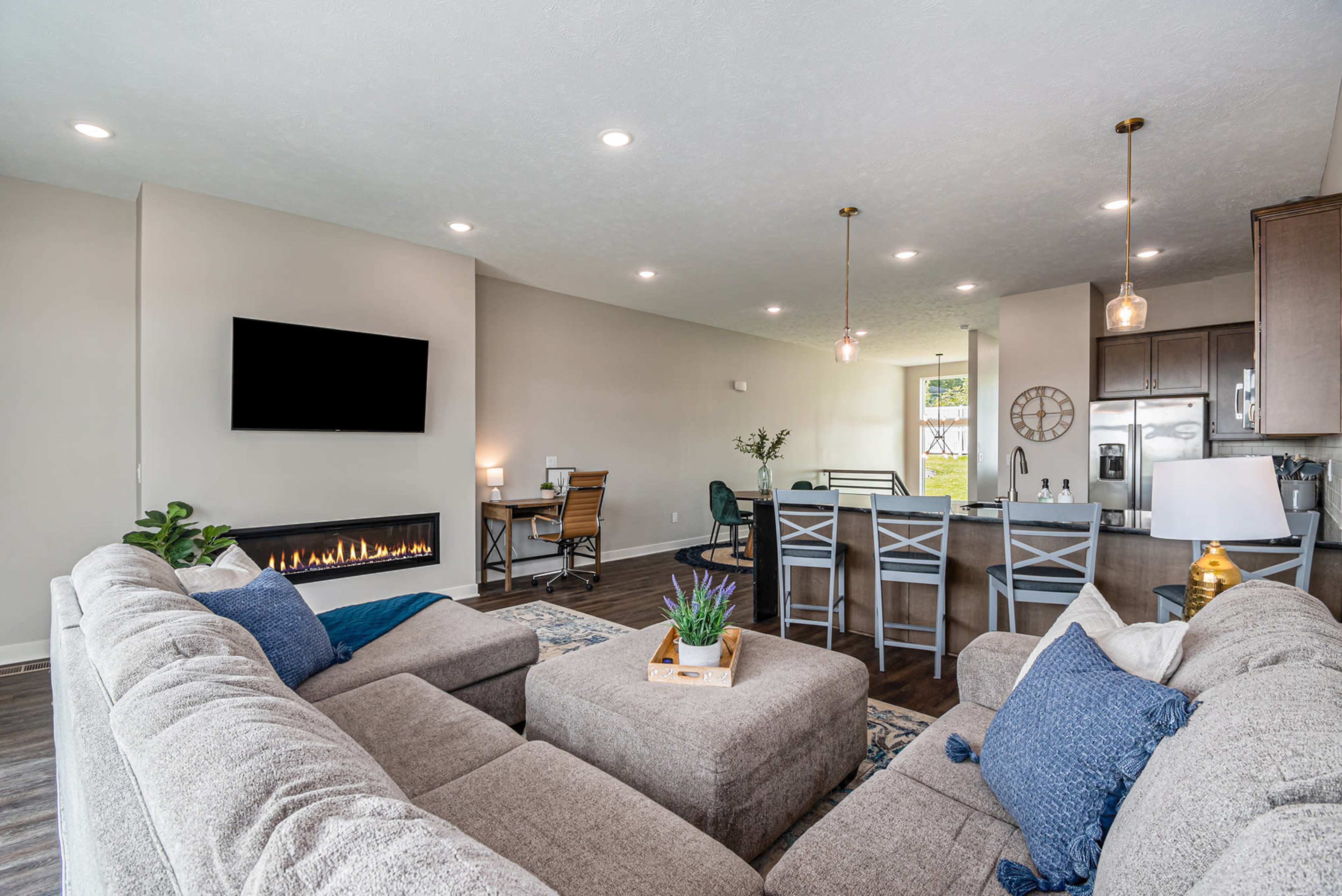 A modern living room features a sectional couch, a flat-screen TV, and a fireplace, with an open layout that connects to a dining area and kitchen.