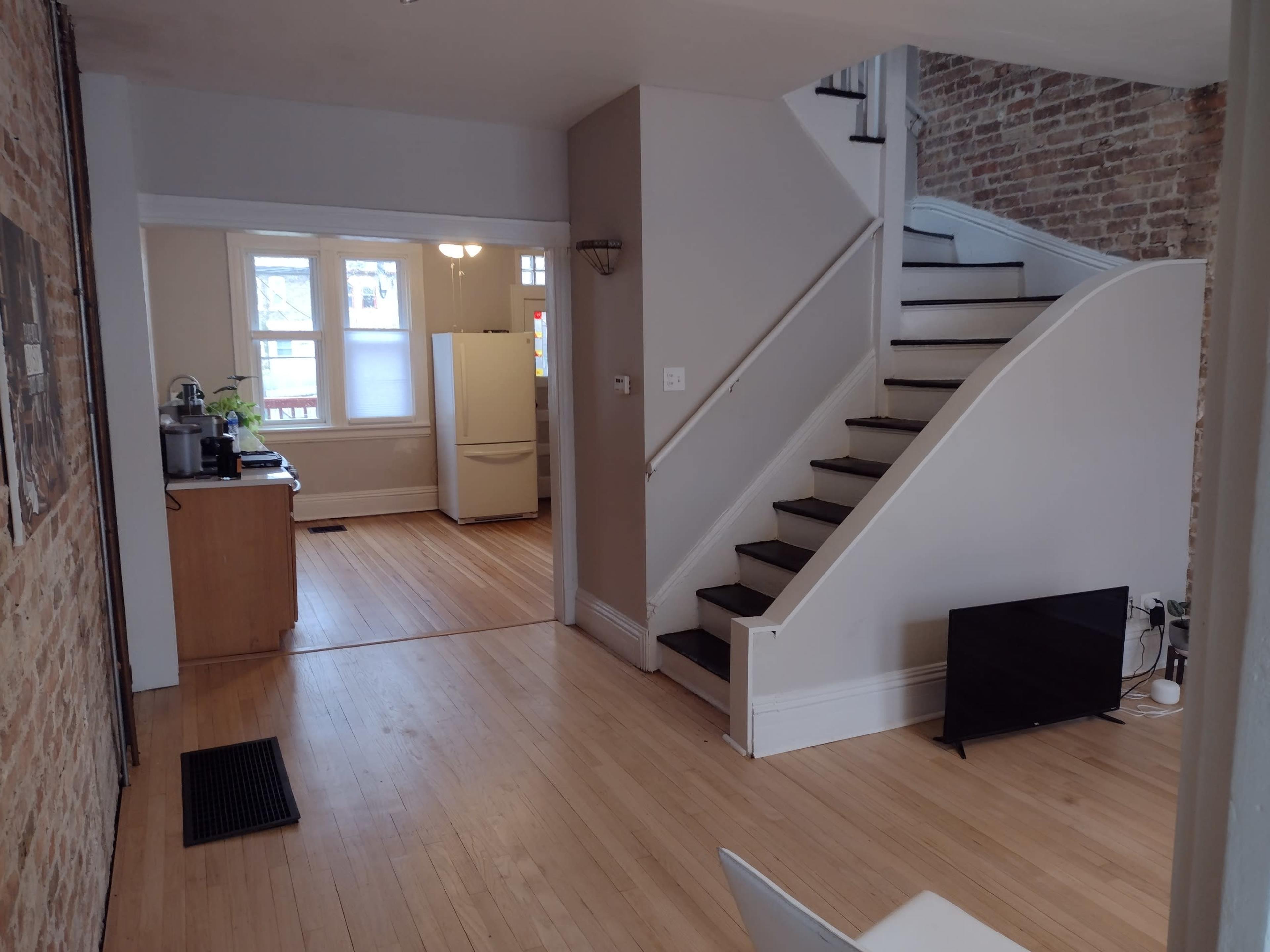A room with hardwood floors, a staircase leading to an upper level, and a view of a kitchen area in the background.