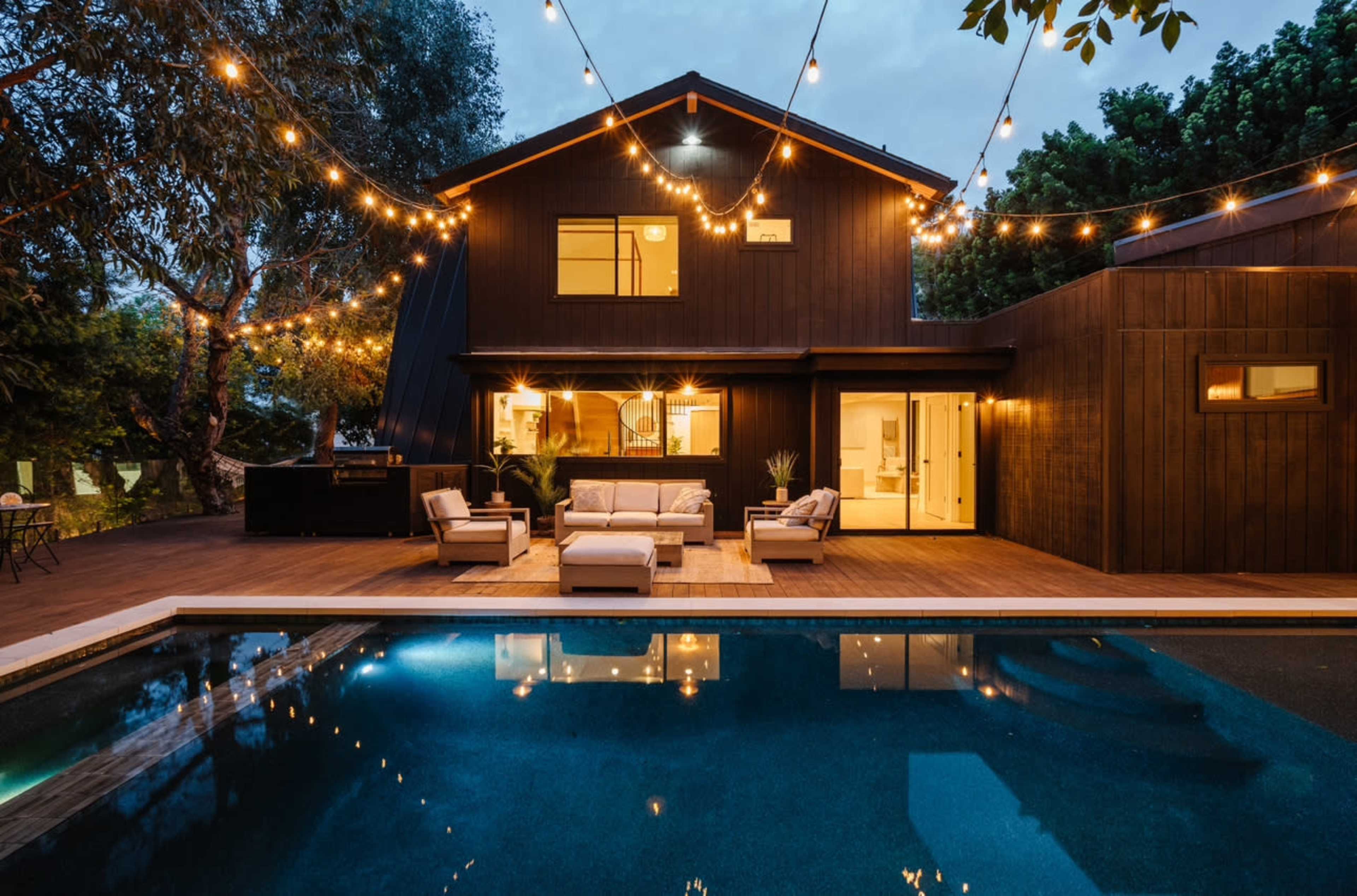 A modern house with a wooden facade is illuminated by string lights, overlooking a swimming pool on a wooden deck.