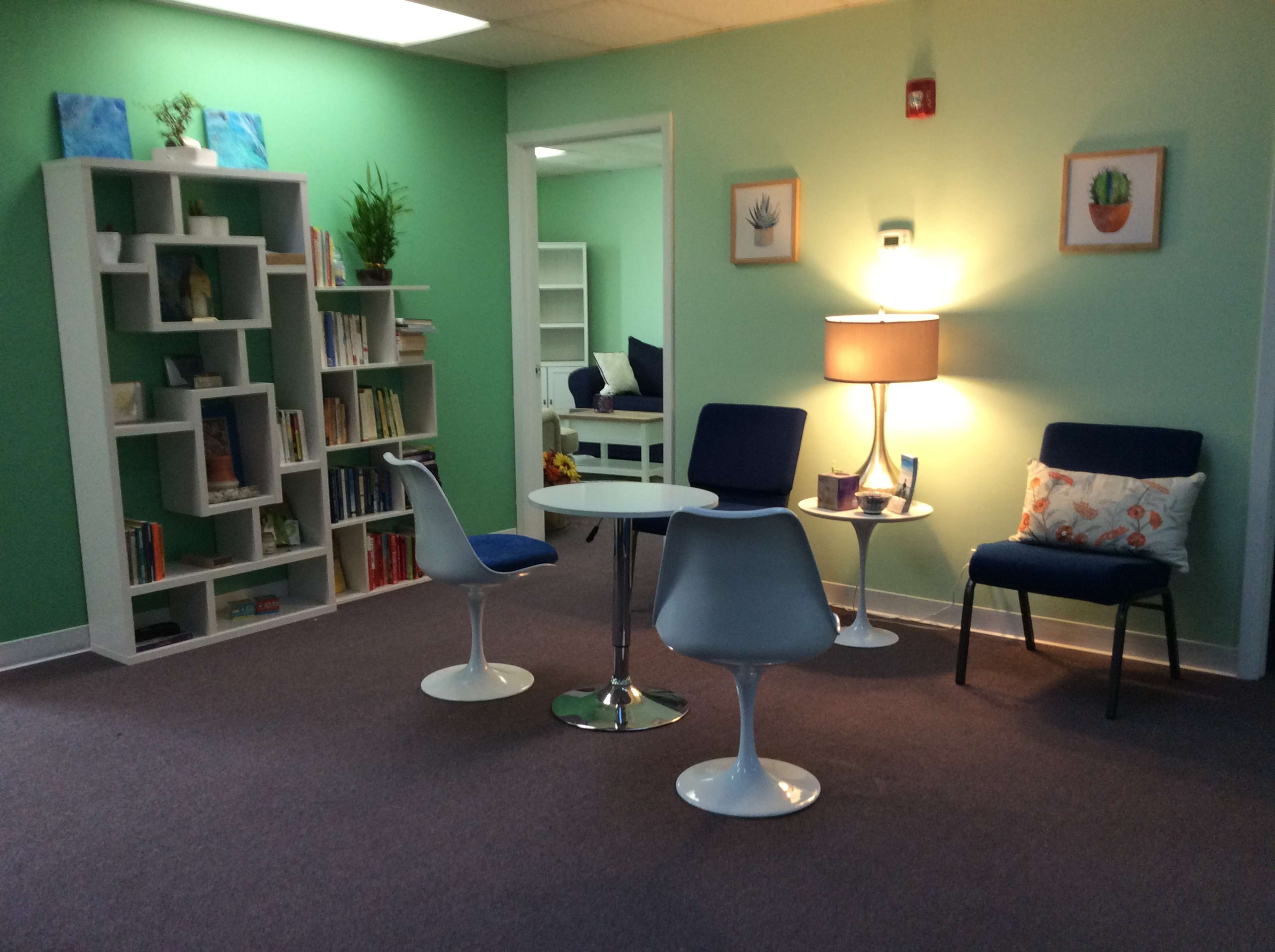A cozy room with green walls, a bookshelf filled with books, a small round table surrounded by white chairs, and a lamp next to a chair.