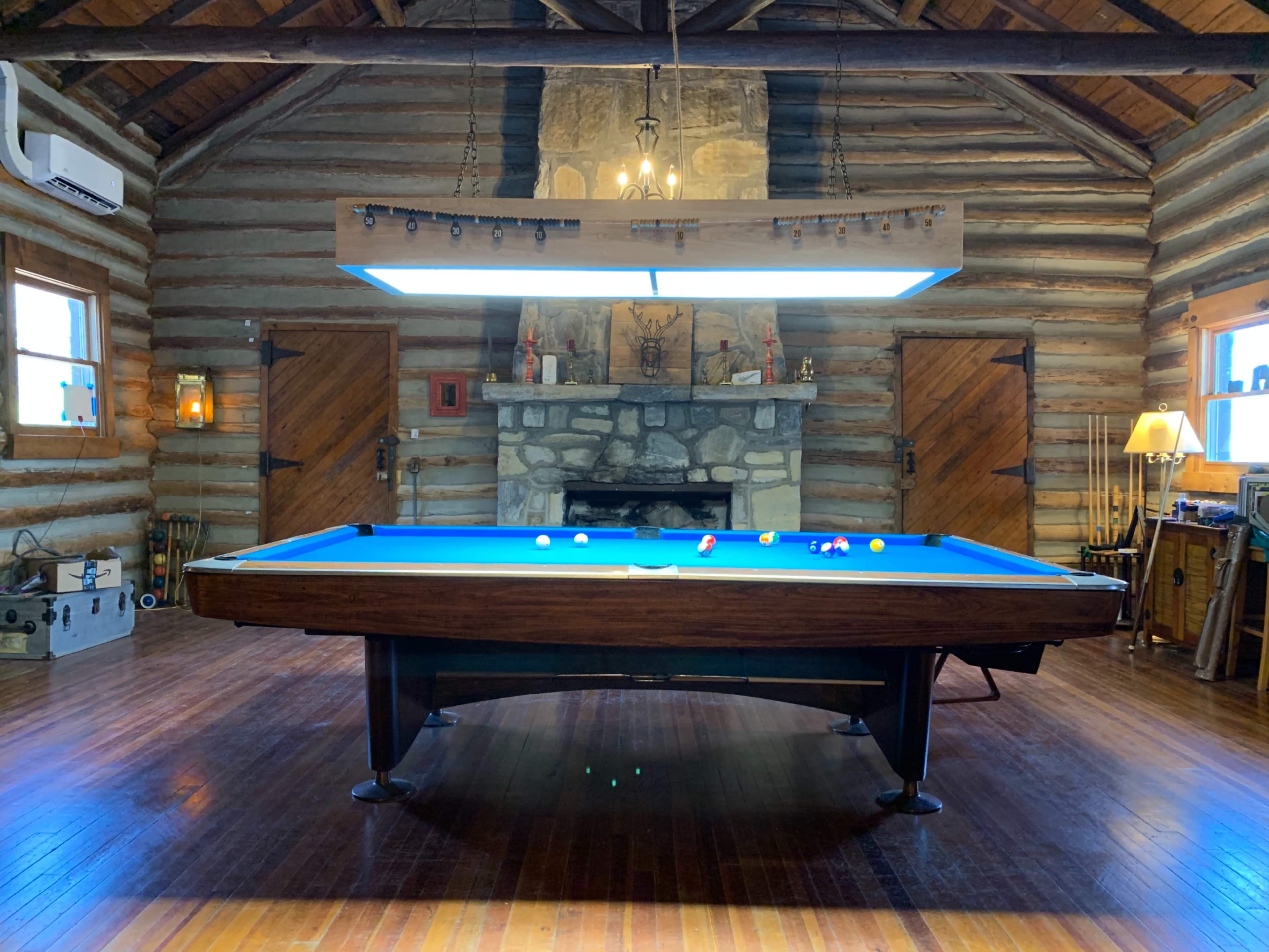 A log cabin interior featuring a blue pool table centered beneath a large overhead light, with a stone fireplace and wooden doors in the background.