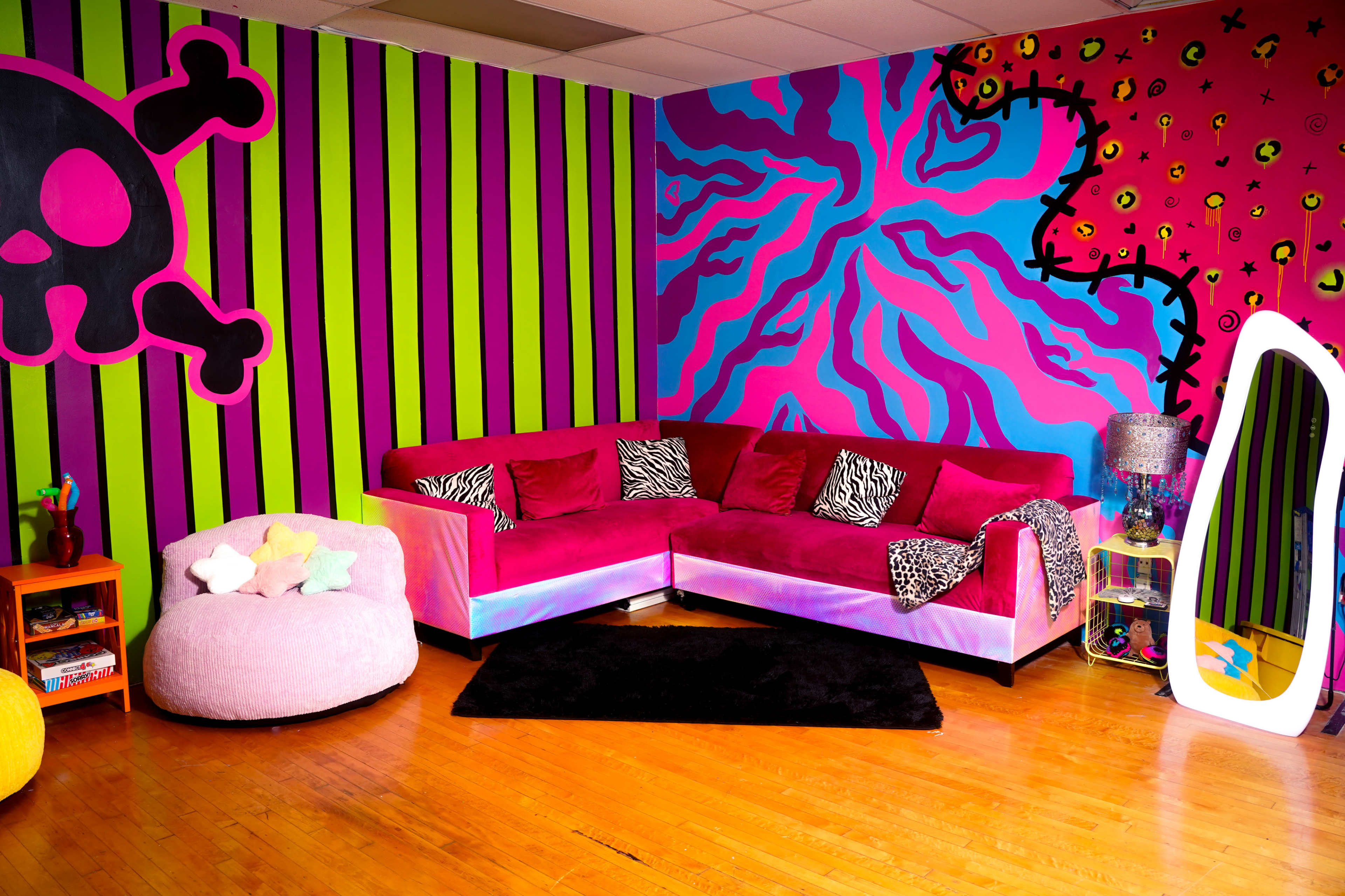The image shows a vibrant room decorated with bold, striped walls in pink and green, featuring a pink sectional sofa, a large bean bag chair, and colorful accessories.