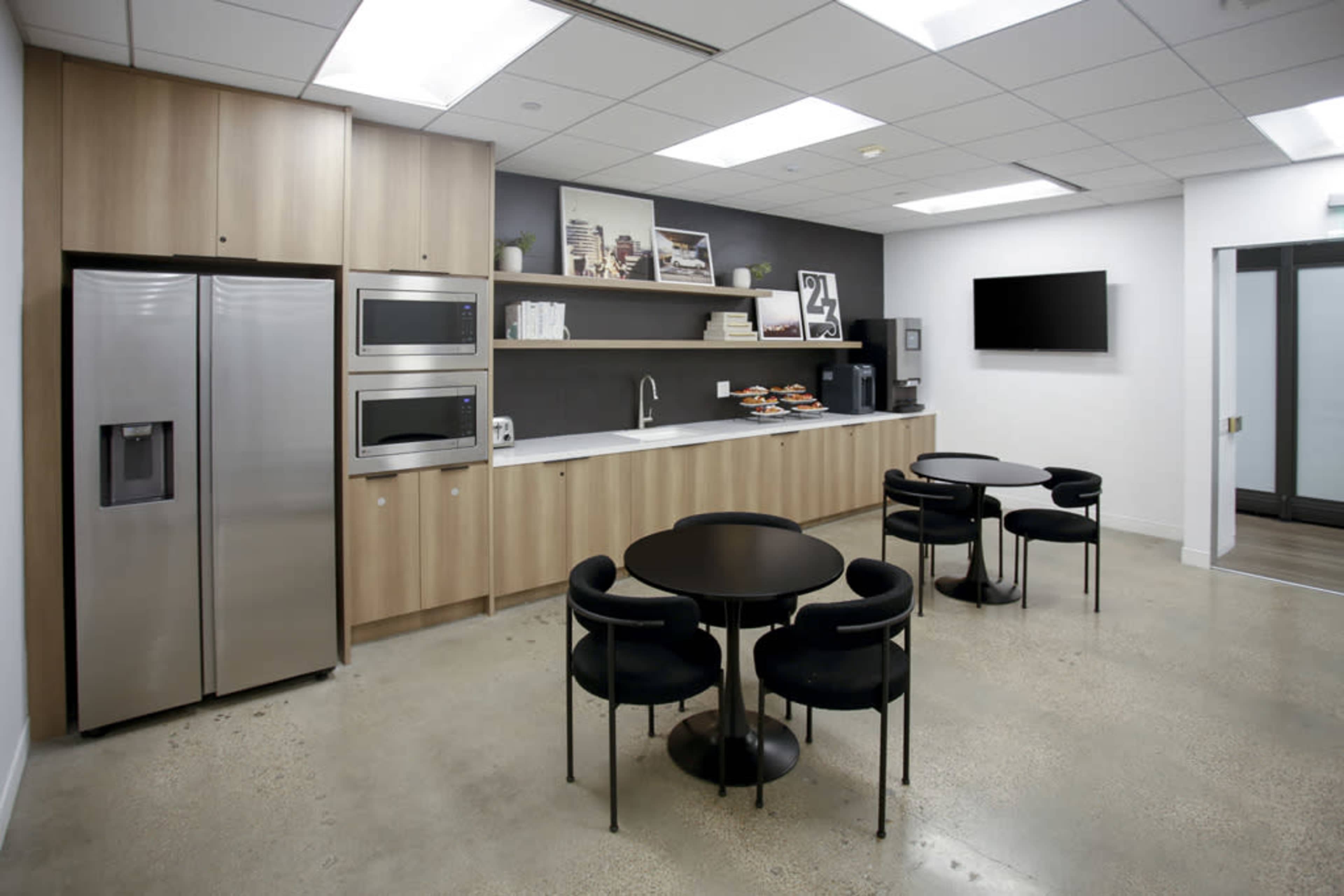 A modern kitchen area with a stainless steel refrigerator, built-in ovens, a long countertop, and round tables with black chairs.