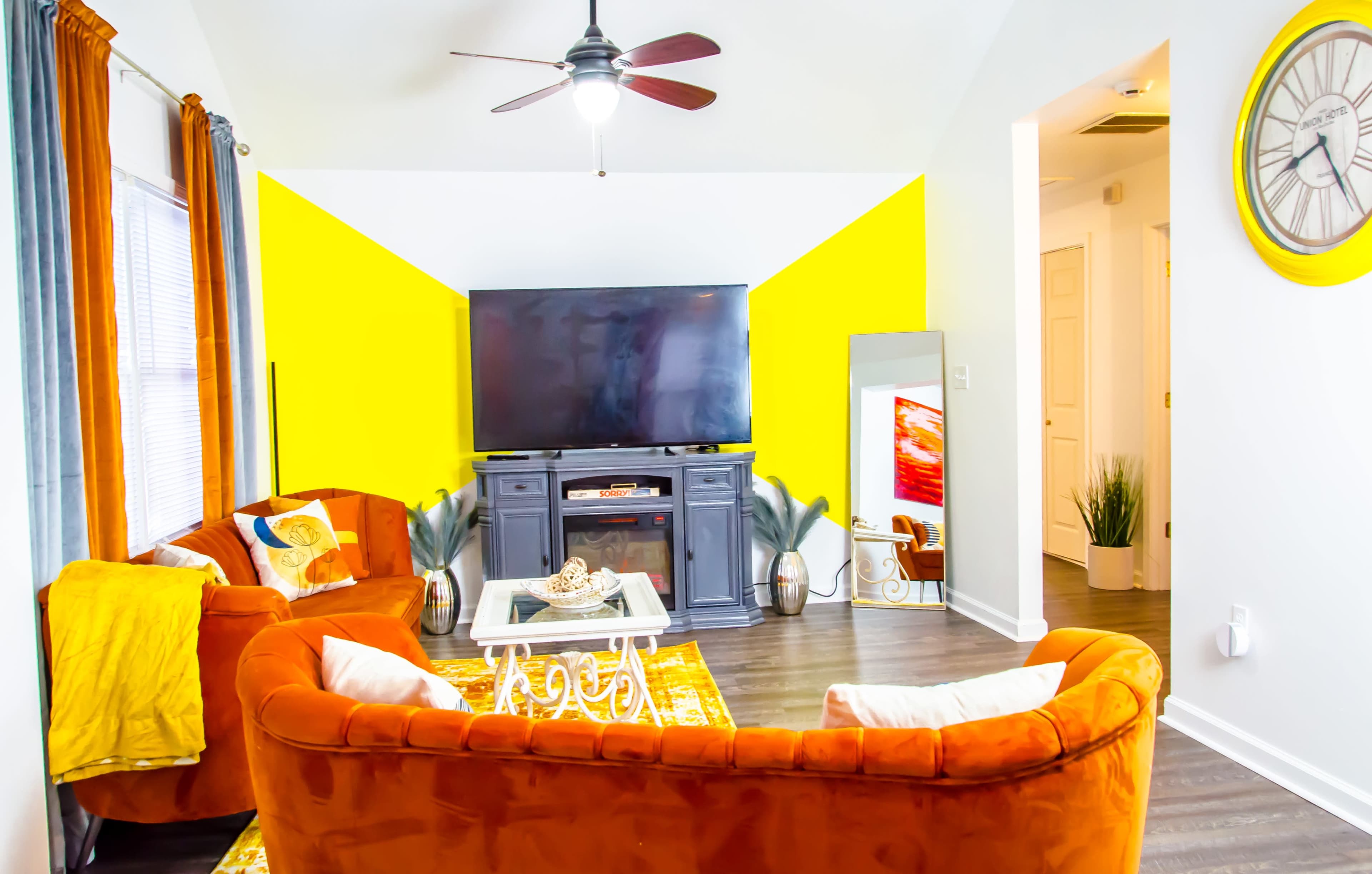 The living room features orange sofas, a large television on a black cabinet, and a bright yellow accent wall.