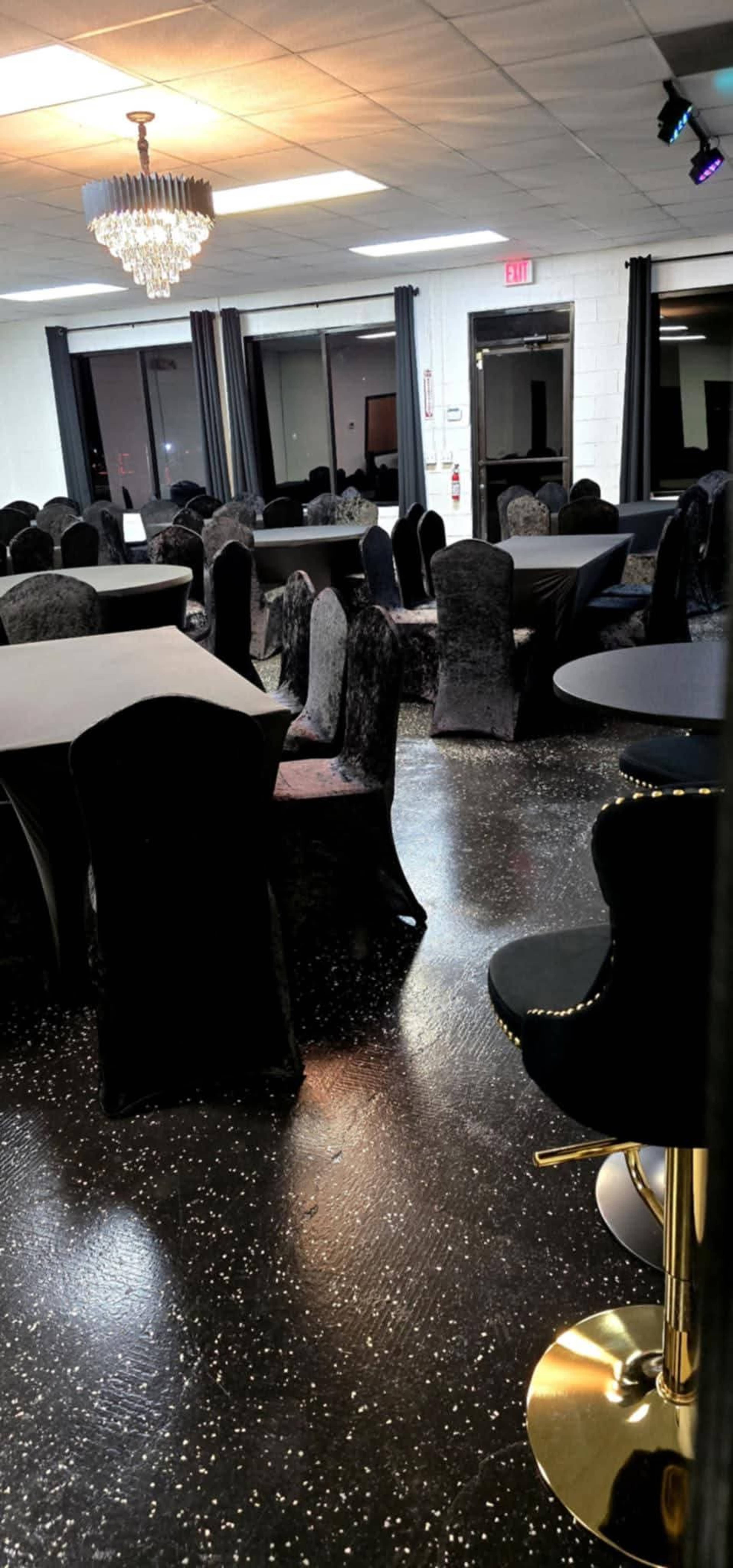 The image shows an empty event space with black tables and chairs, a chandelier hanging from the ceiling, and large windows along one wall.