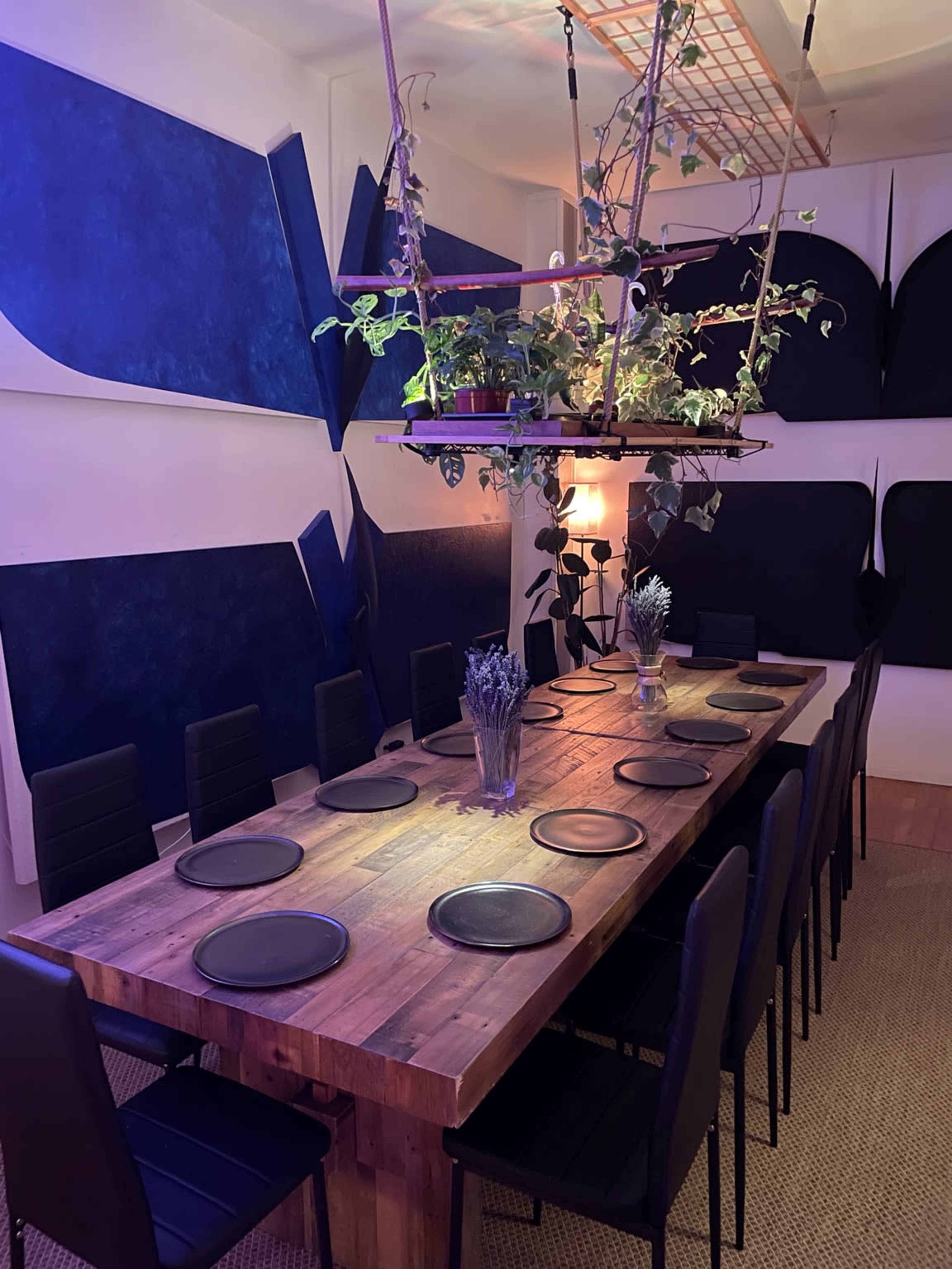 A long wooden dining table is set with black plates and surrounded by black chairs in a room with blue and white acoustic paneling on the walls.